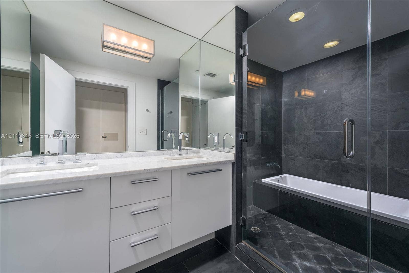 Bathroom featuring white cabinetry, stone-finish vanity countertop, dual undermount sinks with polished chrome fixtures, full-height vanity mirror, and a glass-enclosed bathtub with dark tile surround