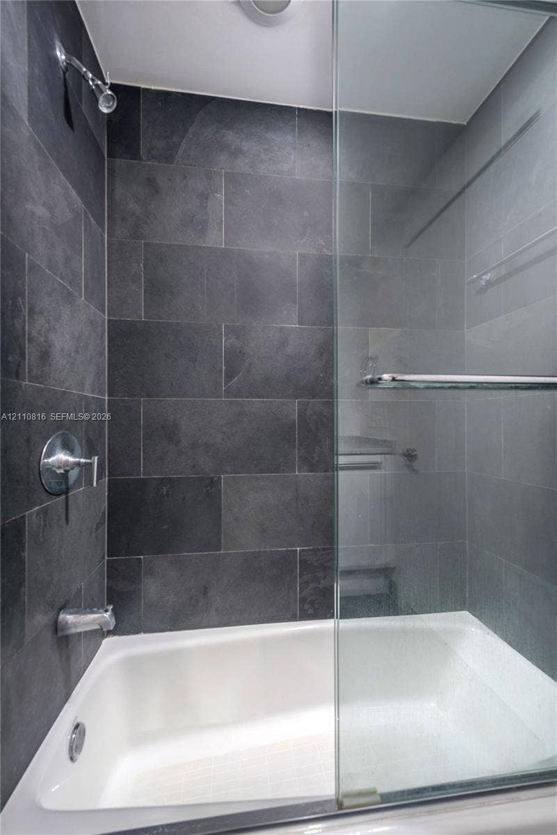 Bathtub with a glass enclosure, featuring large-format dark stone wall tile, chrome shower head, and wall-mounted chrome hardware