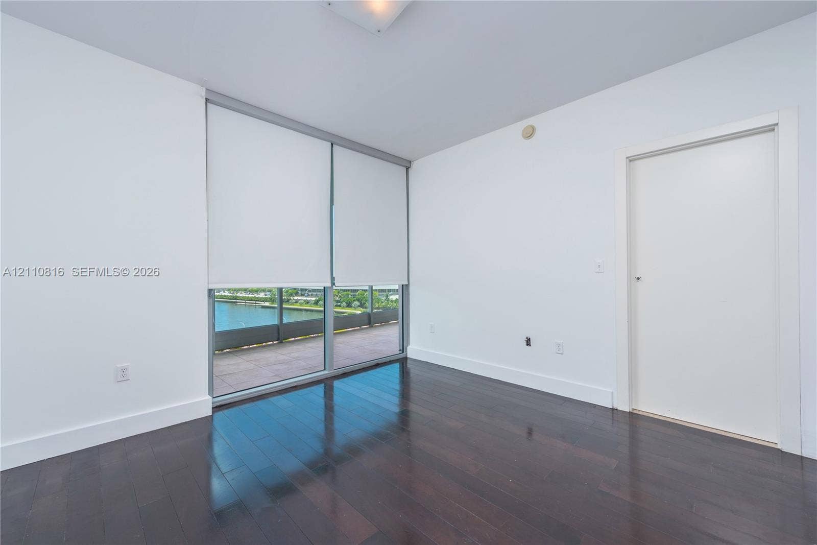 Room featuring dark wood-finish flooring, floor-to-ceiling windows, and access to a balcony with water scenery