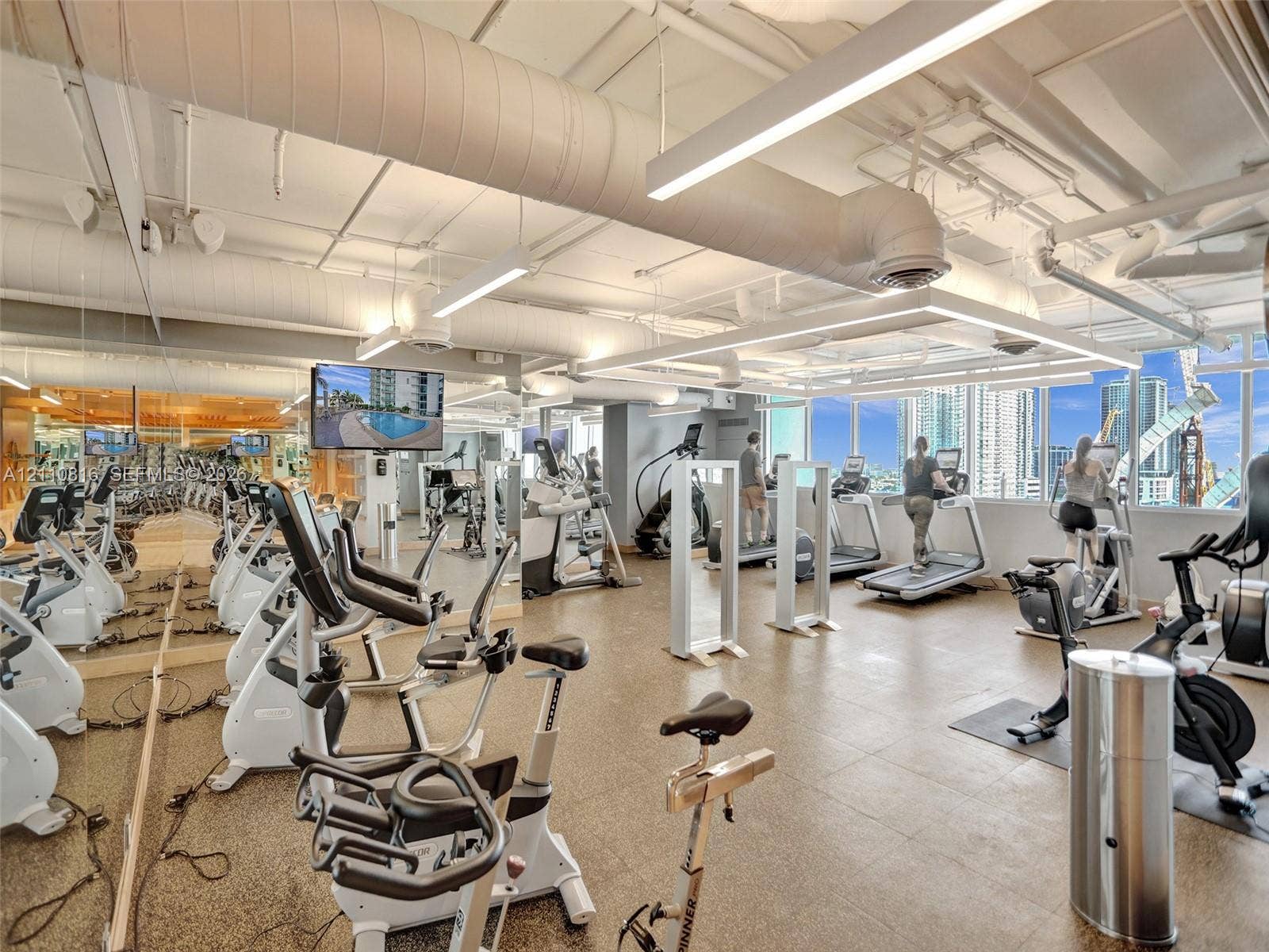 Fitness center featuring industrial-style exposed ductwork, wall-to-wall mirrored surfaces, expansive windows, linear overhead lighting, and stone-finish flooring