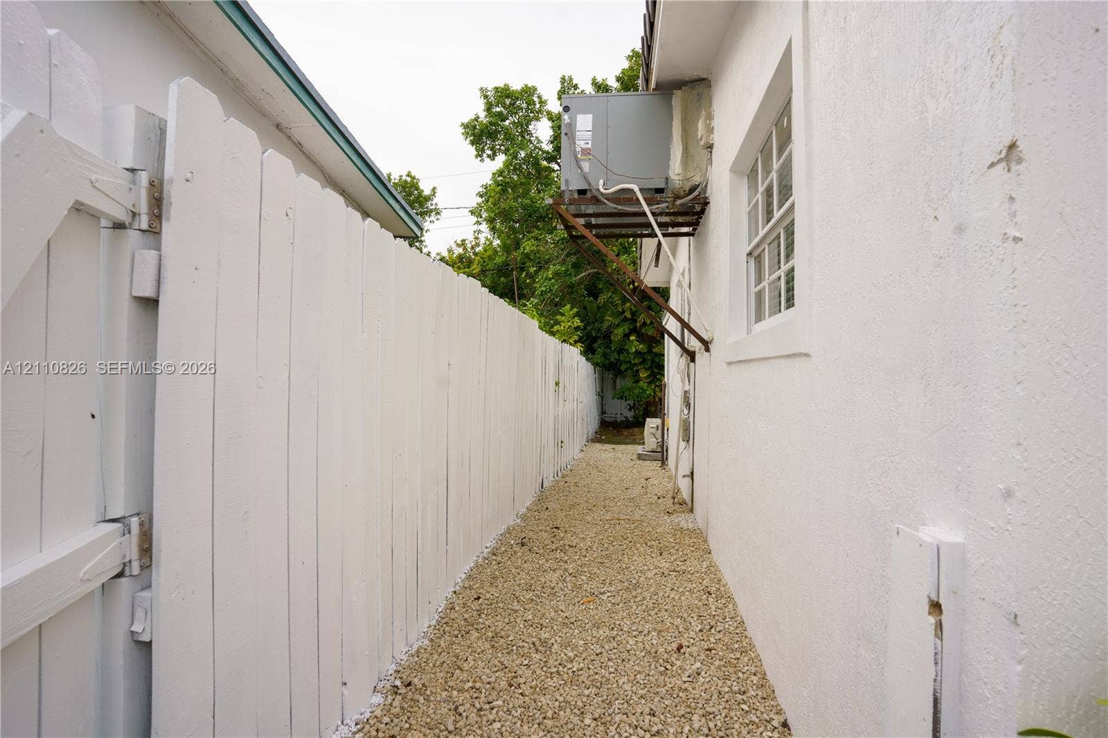 Side exterior area featuring a white picket fence, gravel ground cover, and a wall-mounted HVAC unit