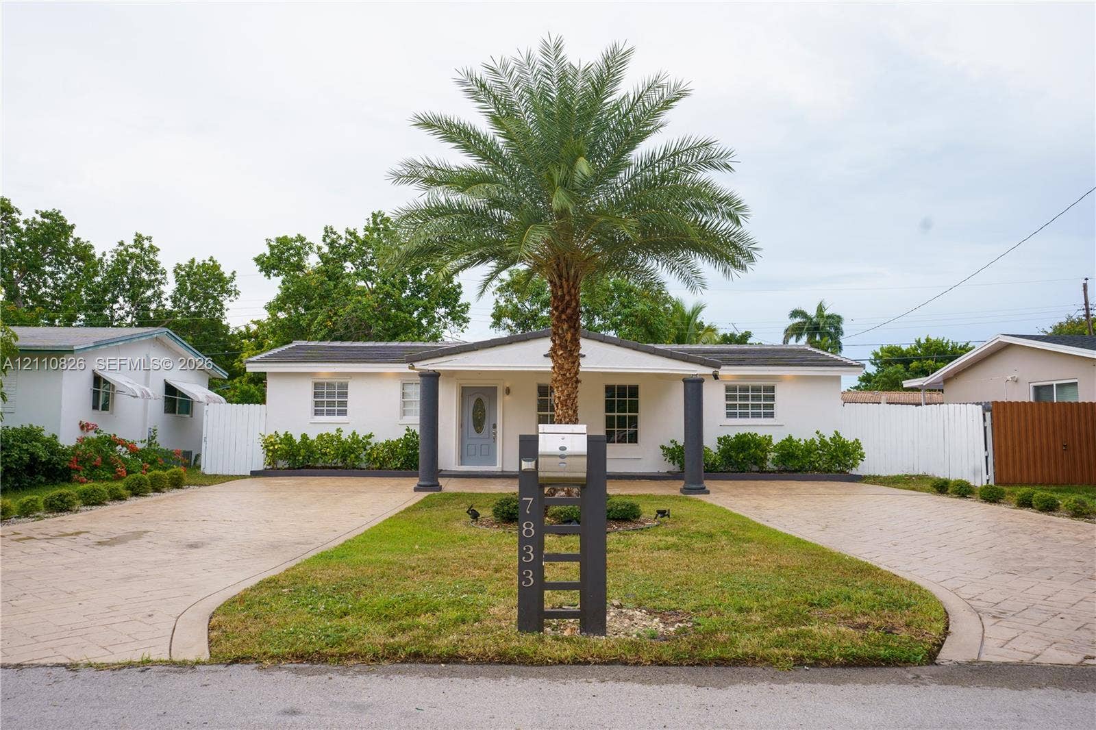 Single-story residence featuring a covered entryway with structural columns, dual paved driveways, a central palm tree, and white privacy fencing