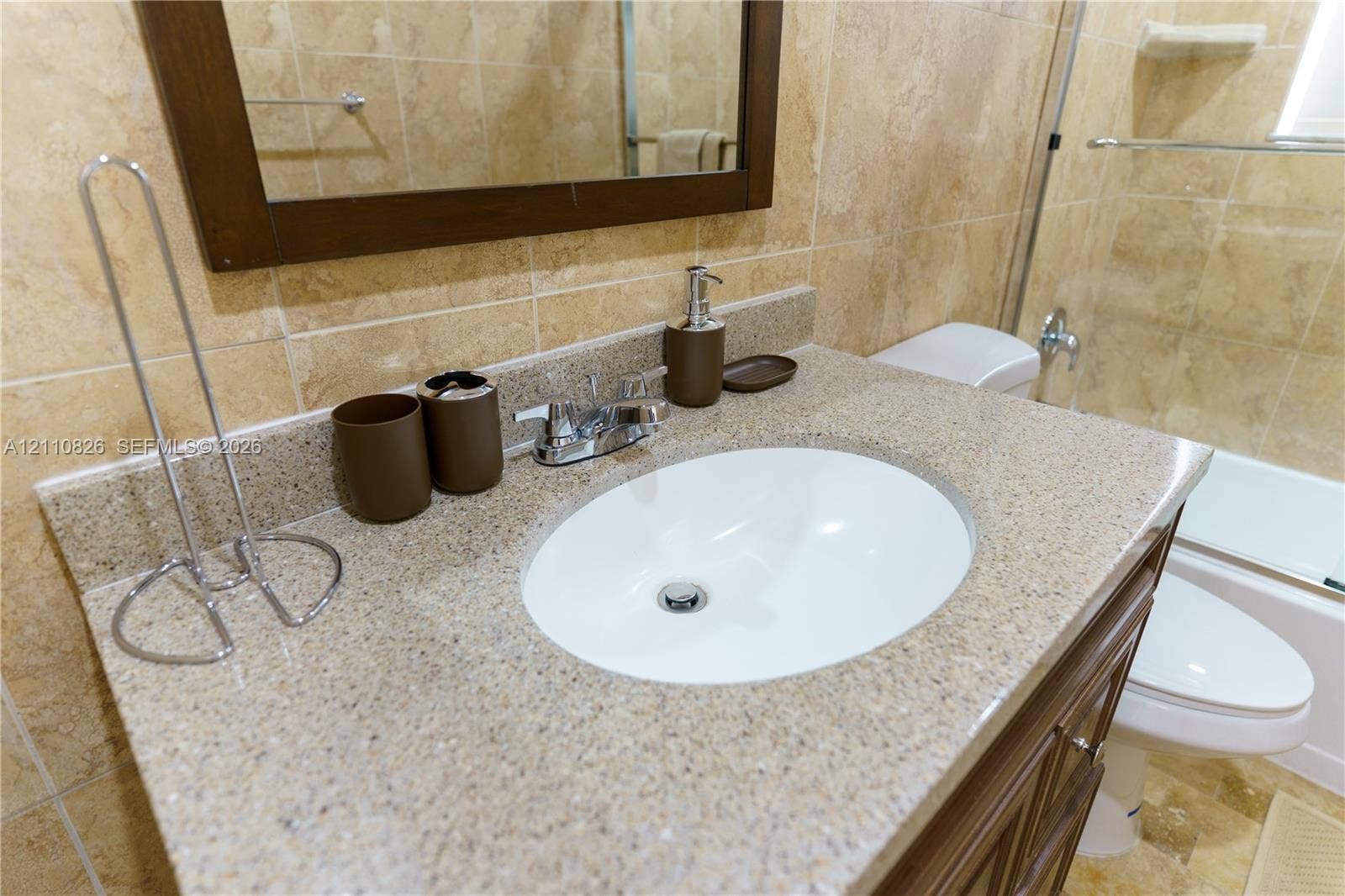 Vanity featuring an integrated oval sink, stone-composite countertop, dark wood-finish cabinetry, and neutral wall tile surround with a glass-enclosed shower