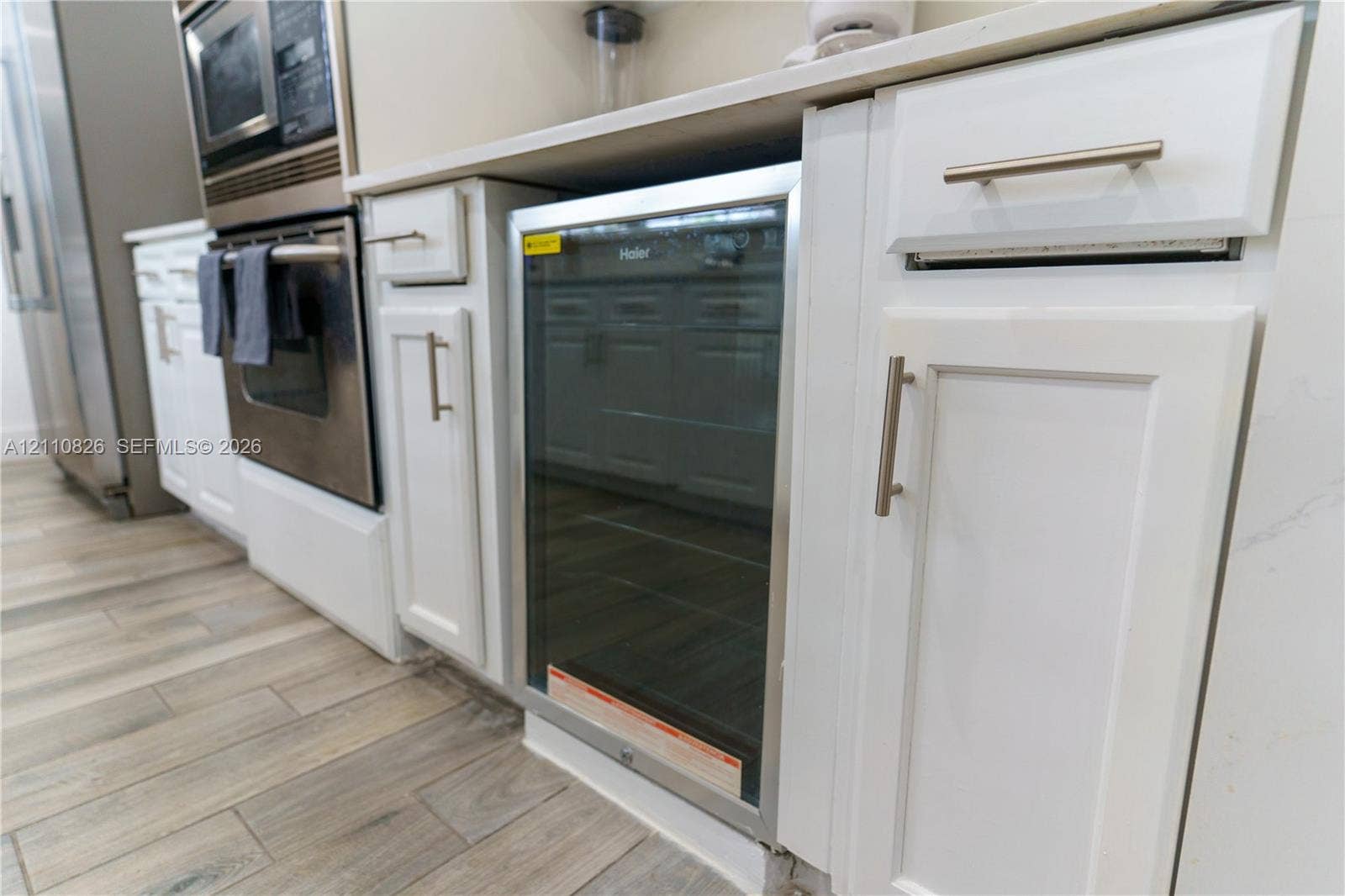 Built-in wine refrigerator, white shaker-style cabinetry with brushed nickel hardware, stone-surface countertops, stainless steel wall oven and microwave, and wood-finish flooring