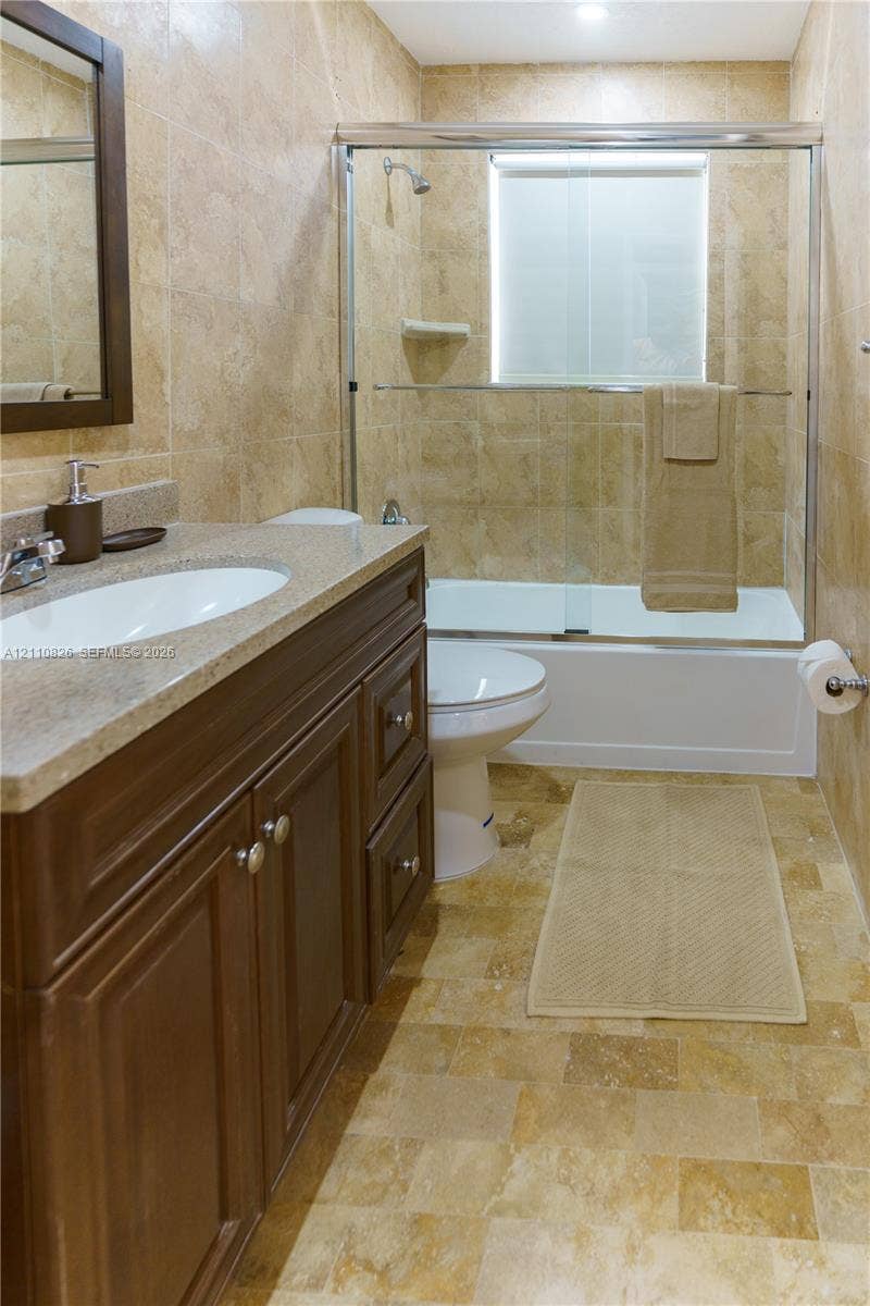 Bathroom featuring a wood-finish vanity with a stone-composite countertop, full-height wall tiling, and a tub-shower combination enclosed by a glass sliding door