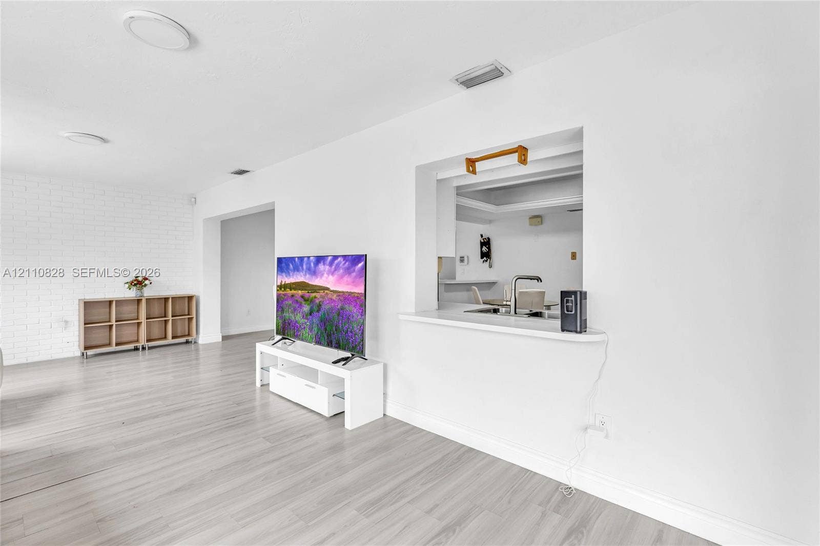 1600 NW 120th Avenue, Pembroke Pines, FL 33026 Interior space featuring light-toned wood-finish flooring, white painted brick accent wall, and a kitchen pass-through window with a countertop ledge