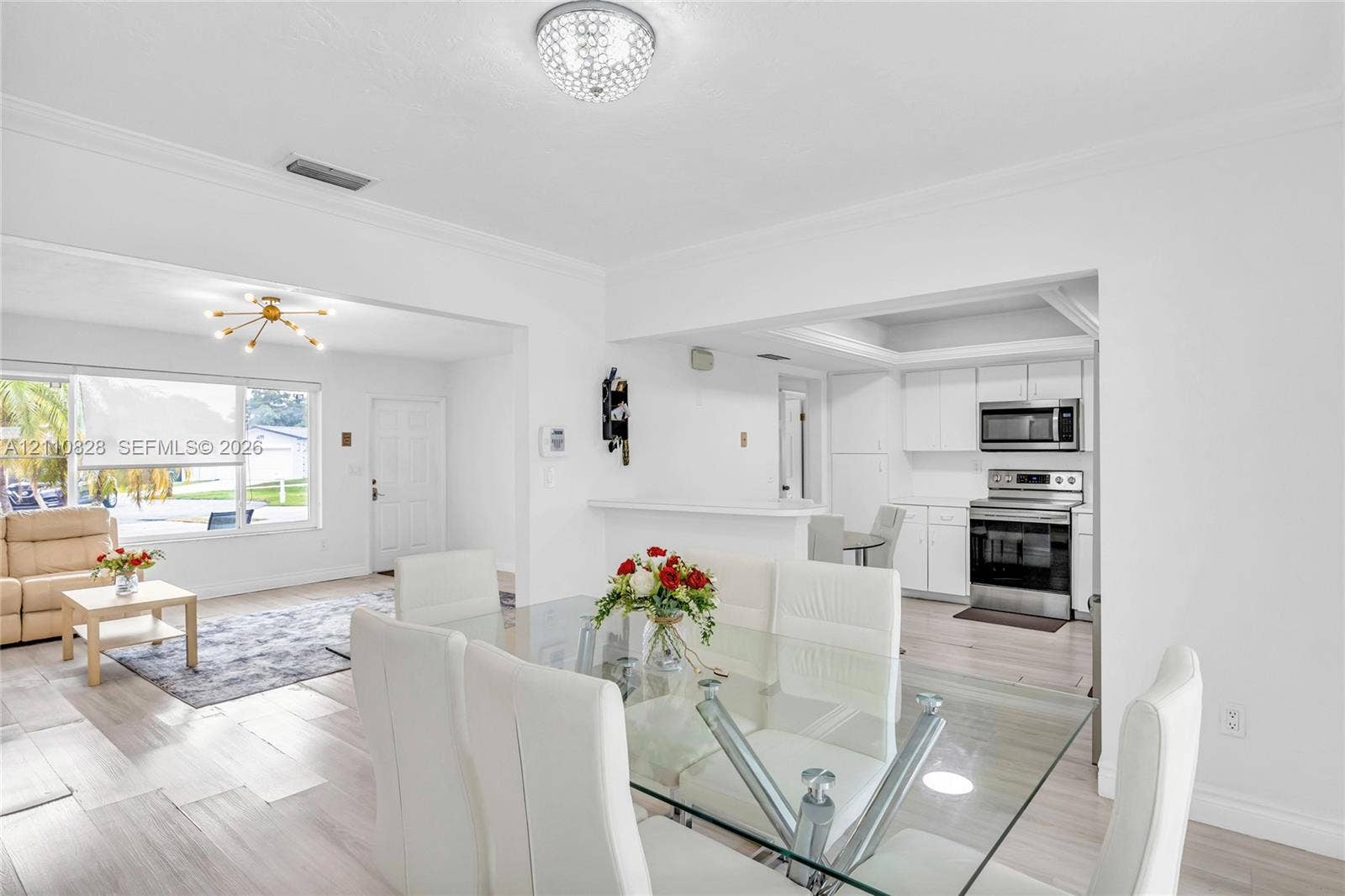 1600 NW 120th Avenue, Pembroke Pines, FL 33026 Open-concept interior featuring light wood-finish flooring, white shaker-style kitchen cabinetry, built-in stainless steel appliances, multi-arm chandelier lighting, and a tray ceiling