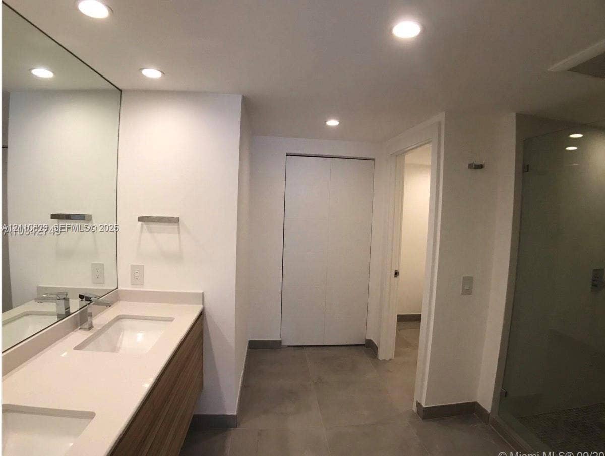 Floating wood-finish double vanity with stone countertop, recessed lighting, expansive wall-to-wall mirror, and glass-enclosed shower with tiled flooring