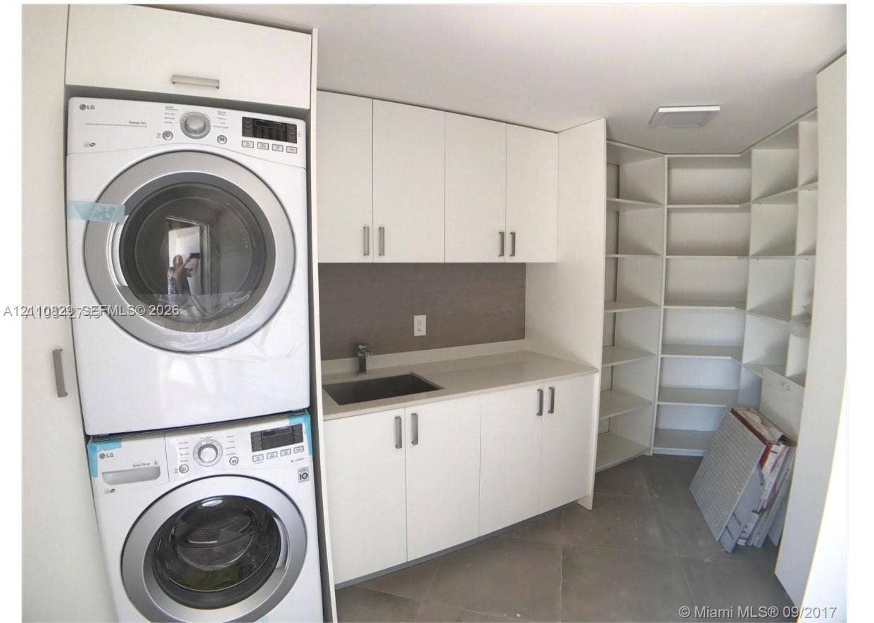 Utility area featuring white flat-panel cabinetry, a solid-surface countertop, an integrated sink with a chrome faucet, a stacked washer and dryer unit, and open built-in shelving