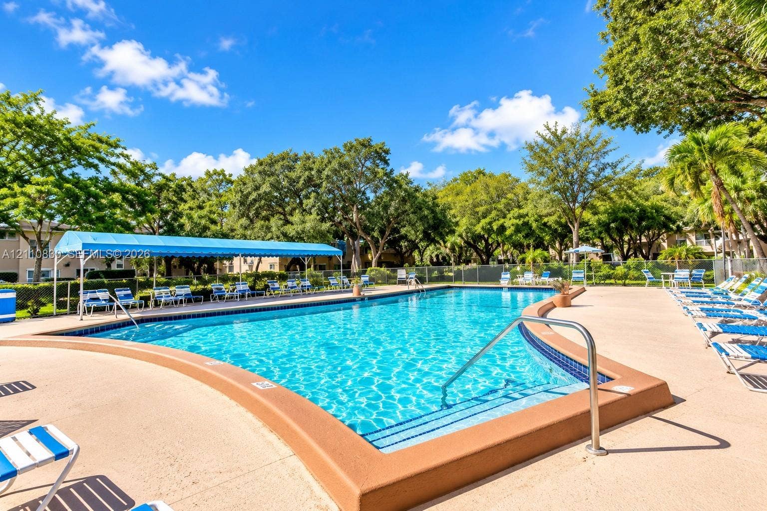 In-ground swimming pool featuring a curved concrete deck, metal entry handrails, blue perimeter tile accents, and a shaded pergola structure