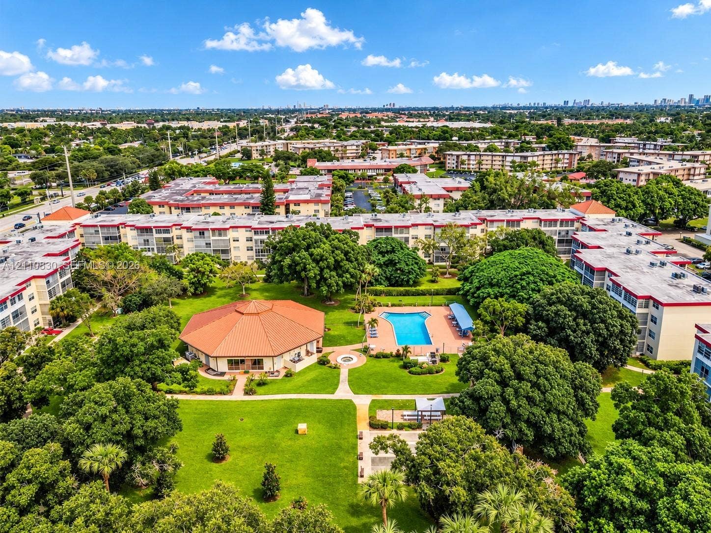 Community grounds featuring an outdoor swimming pool, a clubhouse with a terracotta-style tile roof, mature landscaping, paved pathways, and shaded seating areas