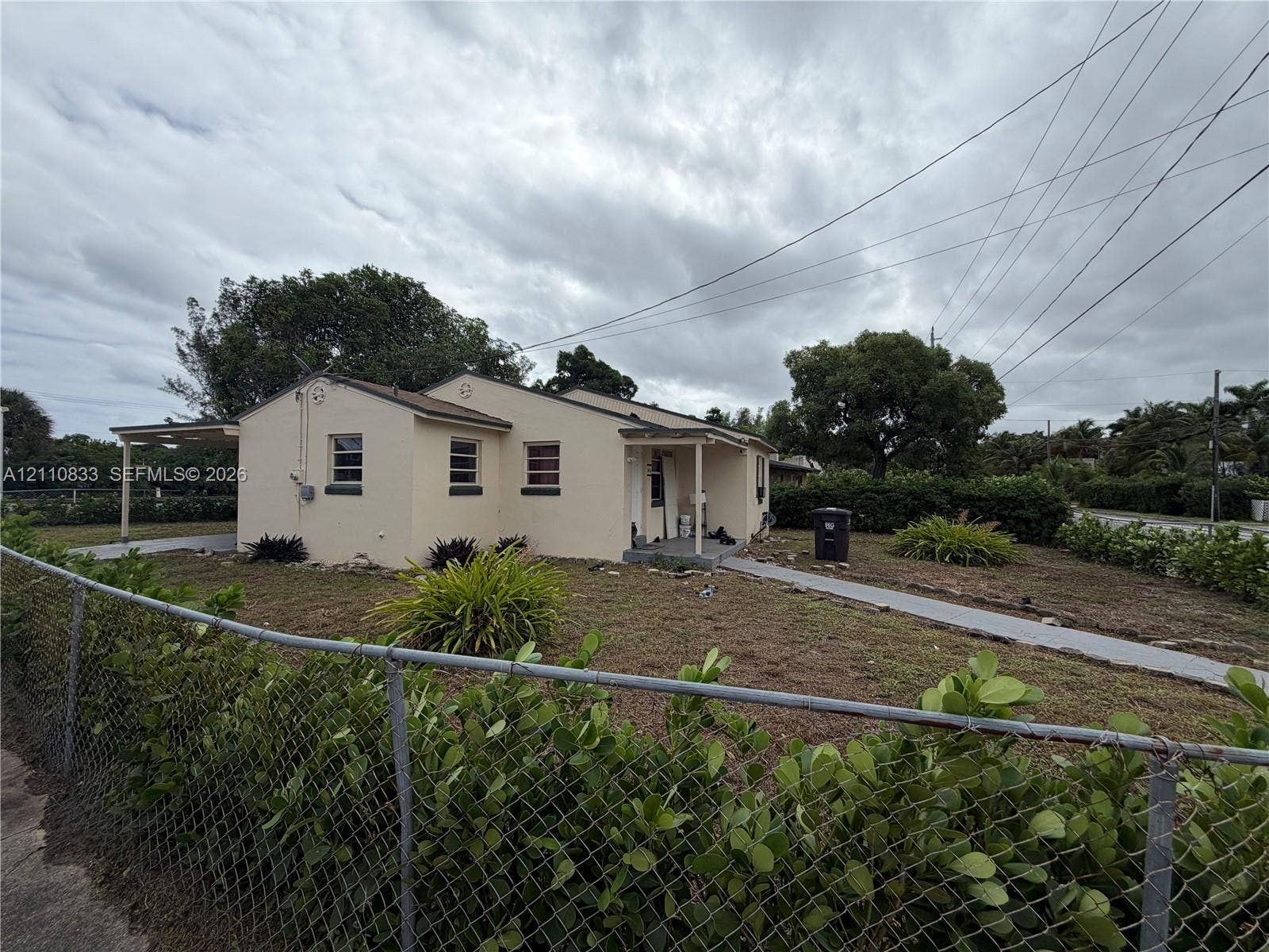 Single-story residence featuring a light-colored stucco exterior, attached carport, concrete walkway, and chain-link perimeter fencing