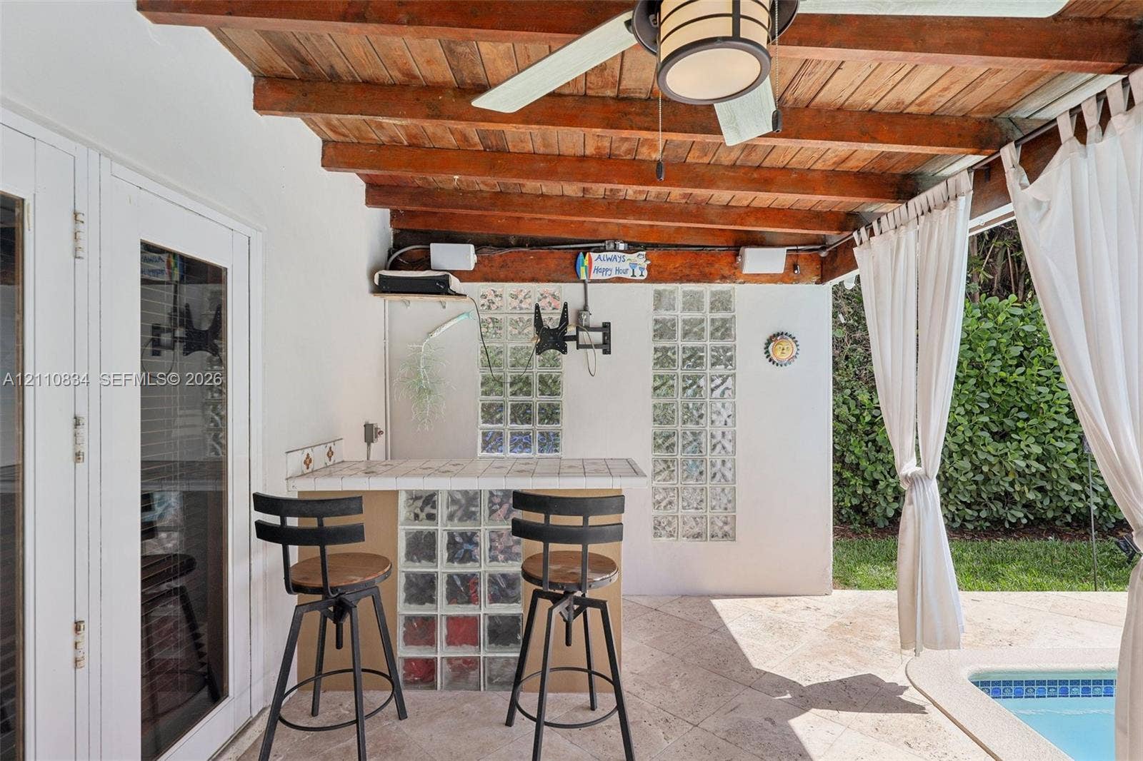 4120 SW 15th Street, Miami, FL 33142 Covered patio featuring an exposed wood-beam ceiling, ceiling fan with integrated lighting, tiled bar with glass-block base, and stone-finish flooring