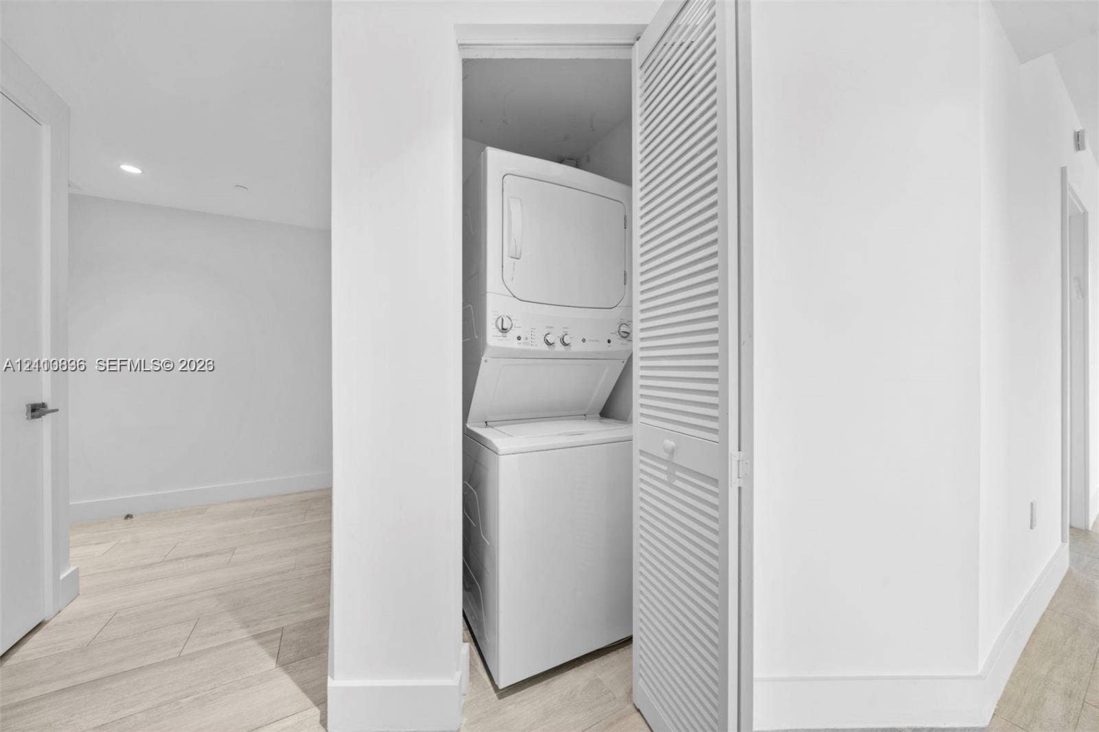 Laundry closet featuring a white stacked washer and dryer unit enclosed by bi-fold louvered doors, situated over light-toned wood-finish flooring