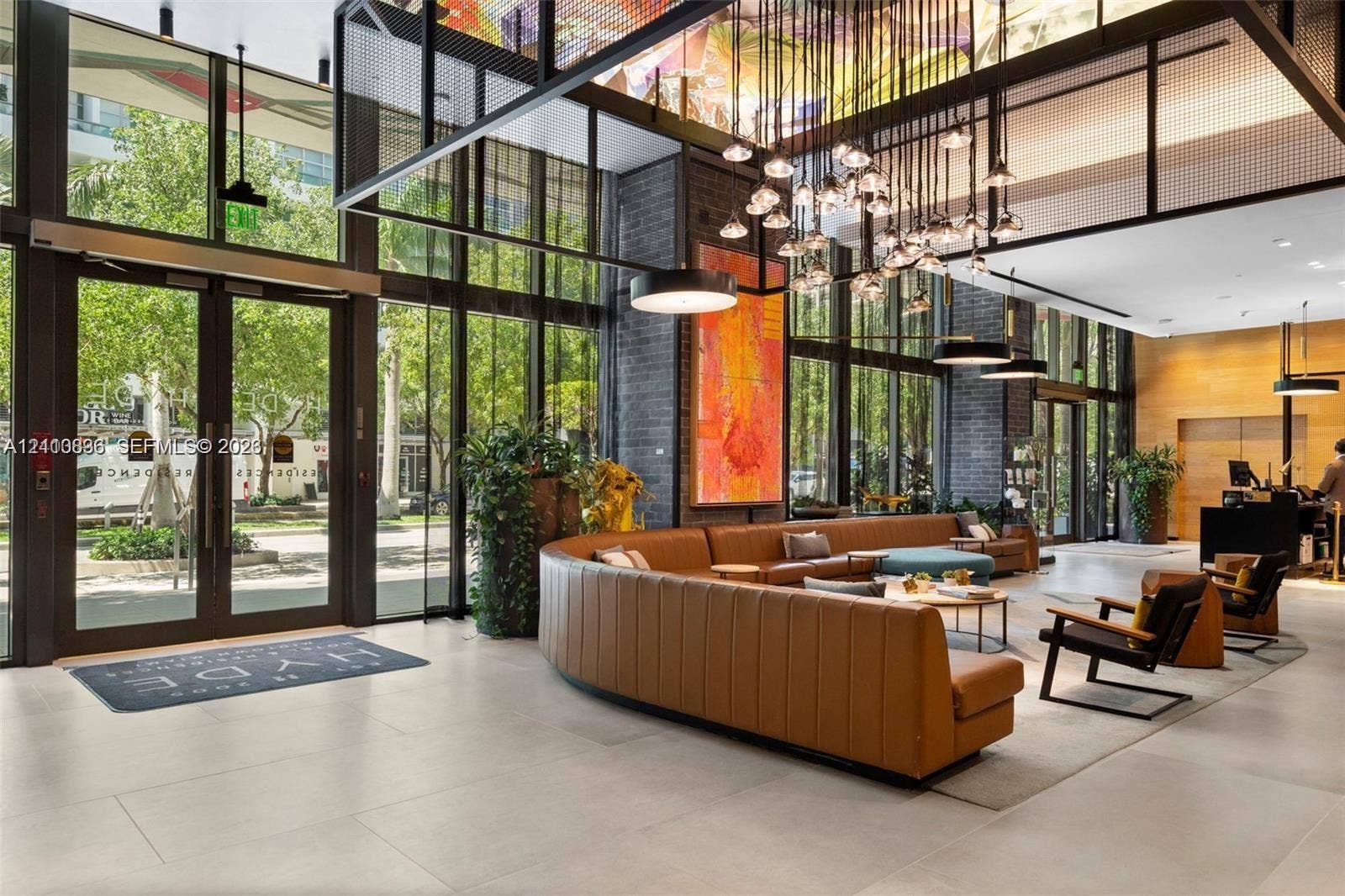 Double-height lobby featuring floor-to-ceiling glass walls, large-format tile flooring, exposed brick structural columns, and a custom multi-light chandelier fixture