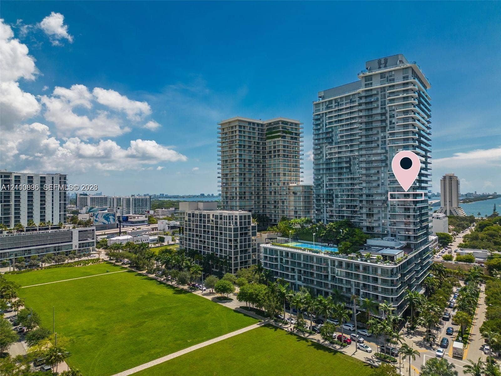 Vibrant urban skyline featuring modern high-rise residential towers with glass and metal facades, manicured green space with mature landscaping, tree-lined streets, and waterfront proximity visible in the distance