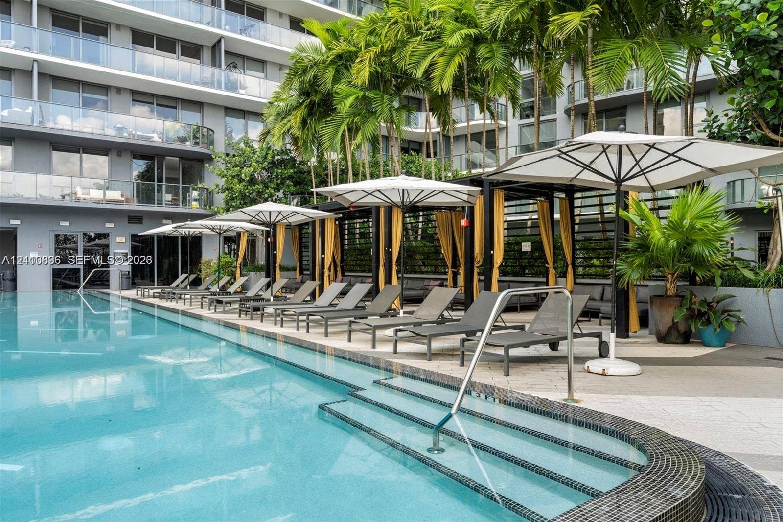 Resort-style pool deck featuring a sparkling lap pool with lane dividers, multiple white umbrellas with integrated shade structures, lounge seating, lush tropical landscaping with mature palm trees, and modern multi-story residential building backdrop