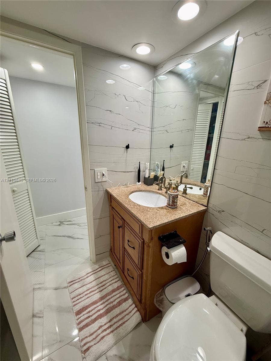 565 Oaks Lane, #410, Pompano Beach, FL 33069 Full-height wall tiling featuring stone-patterned surfacing, vanity with integrated stone countertop and oval undermount sink, wall-spanning vanity mirror, recessed ceiling lighting, and large-format floor tiling
