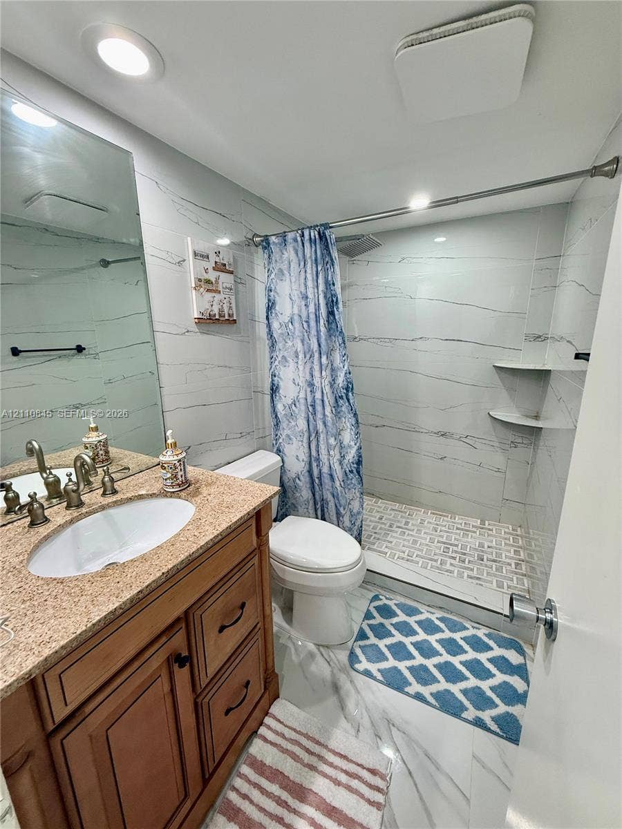 565 Oaks Lane, #410, Pompano Beach, FL 33069 Bathroom featuring a wood-finish vanity with a stone-composite countertop, undermount sink, recessed lighting, and a walk-in shower with stone-patterned wall tile and built-in shelving