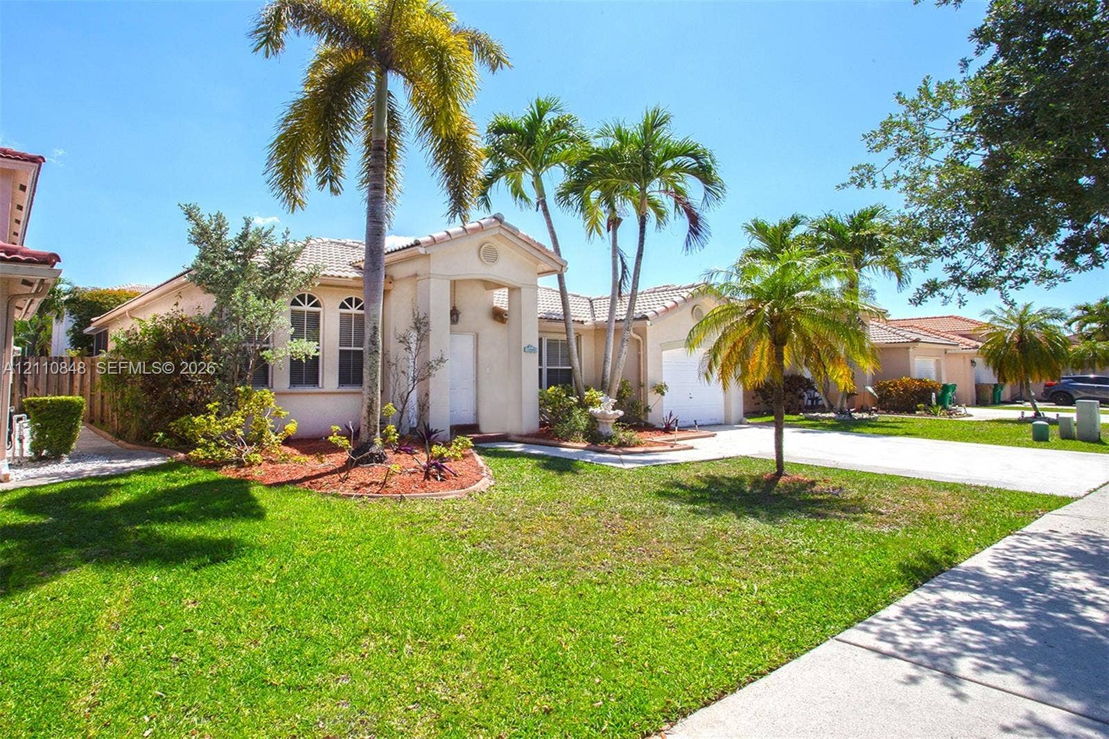 Stucco exterior residence featuring a tile roof, arched windows, covered entryway, and an attached two-car garage set on a landscaped lot with mature palm trees