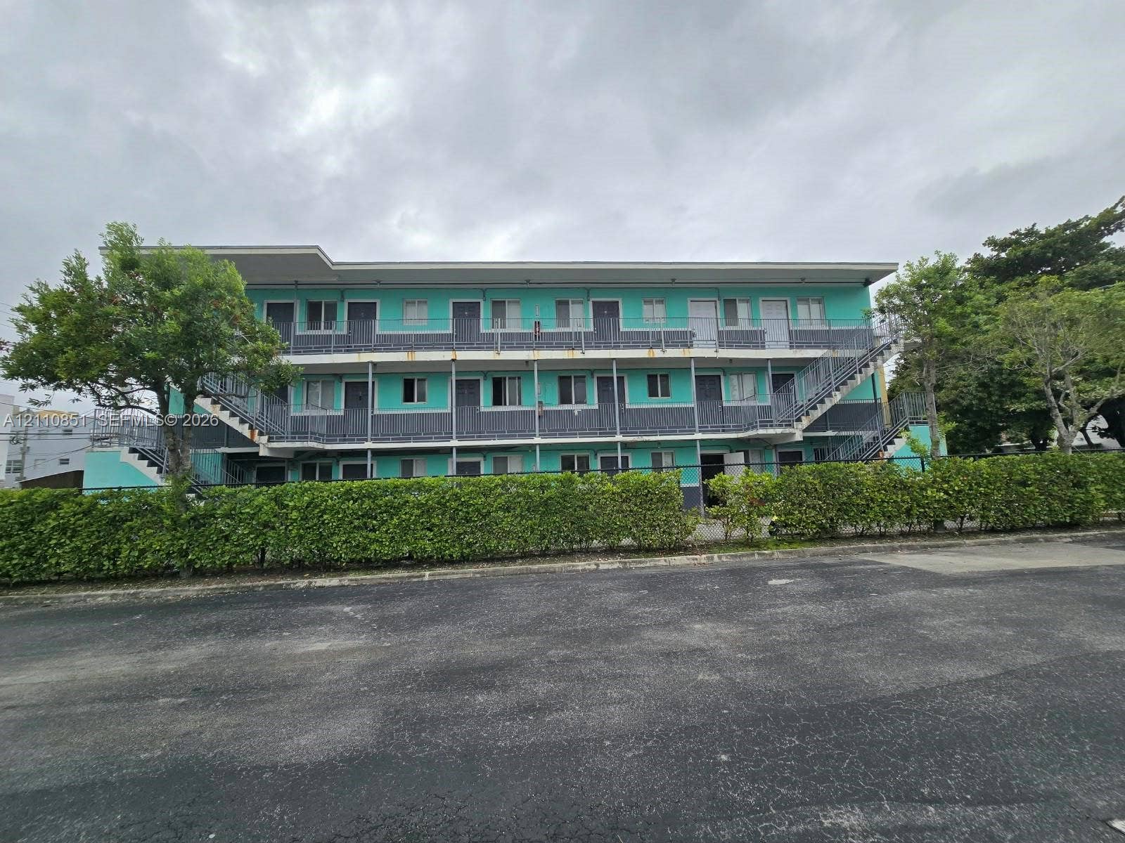 Multi-story building exterior featuring a teal and gray facade, metal staircases, exterior walkways, and mature landscaping