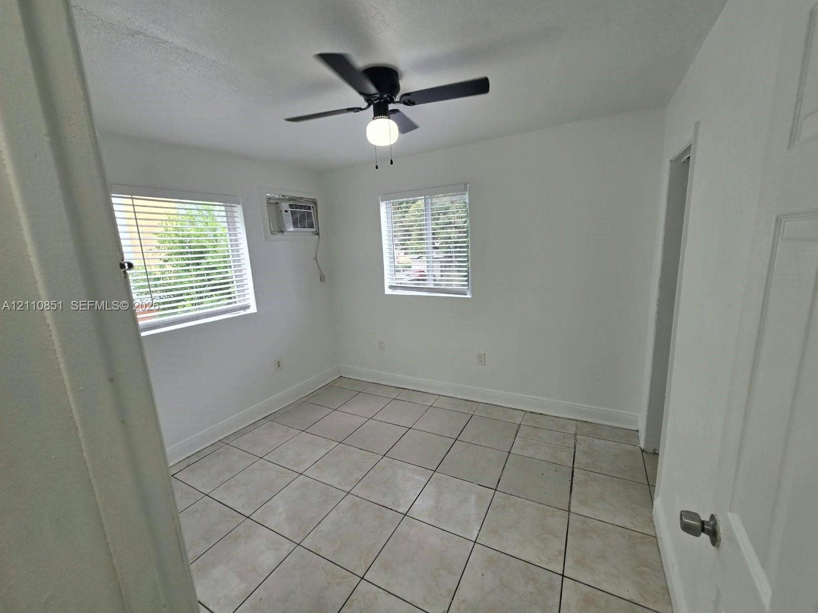 Room featuring neutral tile flooring, dual windows with horizontal blinds, and a ceiling-mounted light fixture with fan