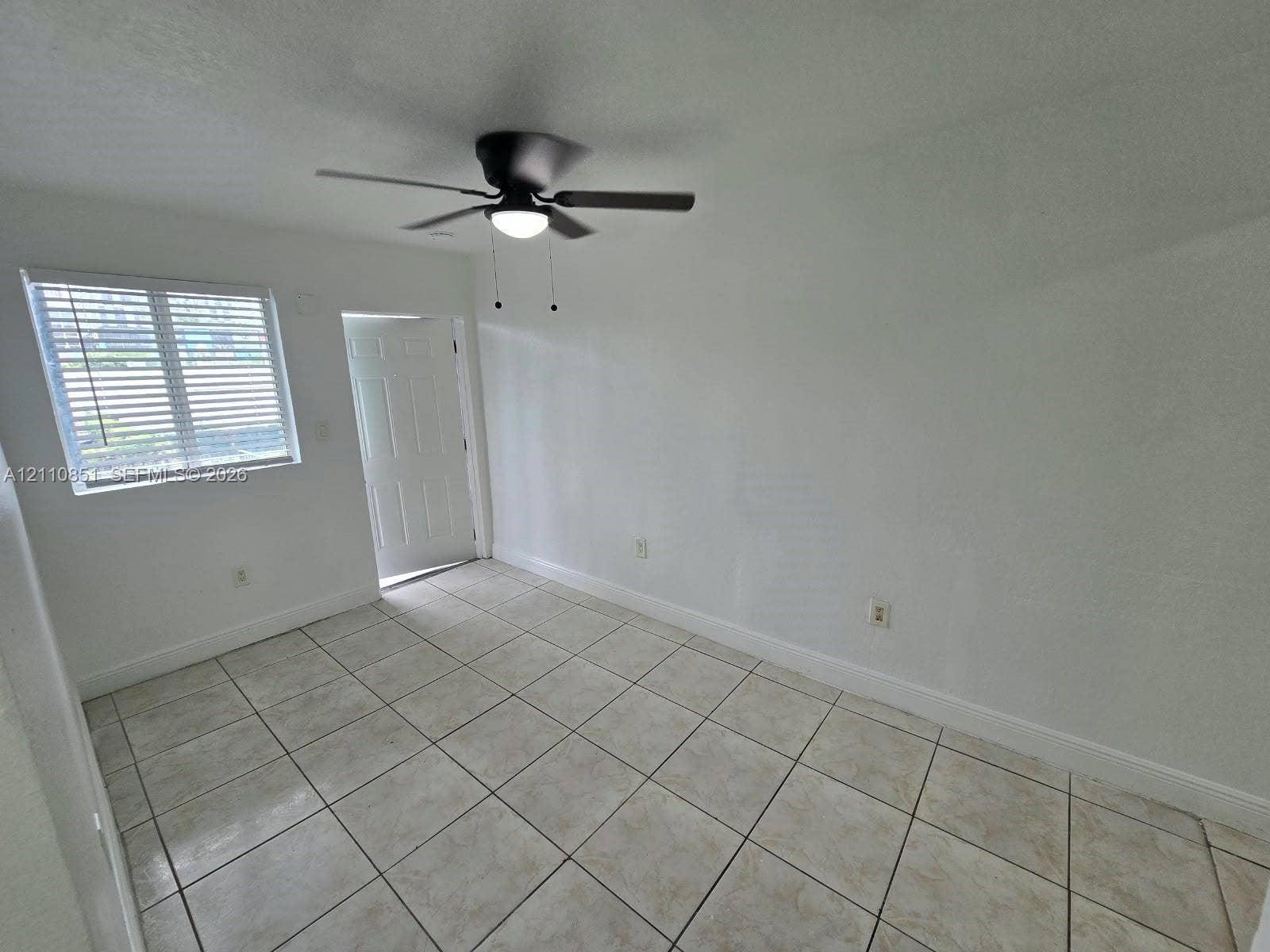 Neutral-toned tile flooring, multi-panel door, single-hung window with horizontal blinds, and a matte black ceiling fan with integrated light fixture