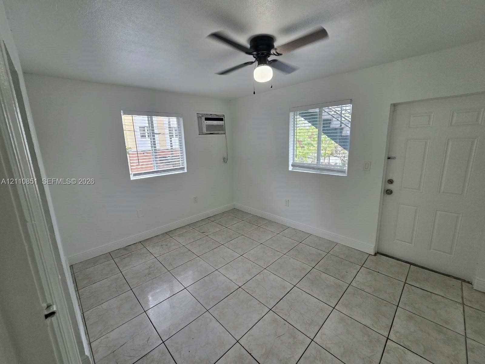 Neutral-toned tile flooring, white wall finishes, two windows with horizontal blinds, built-in wall-mounted air conditioning unit, and a ceiling fan with integrated light fixture