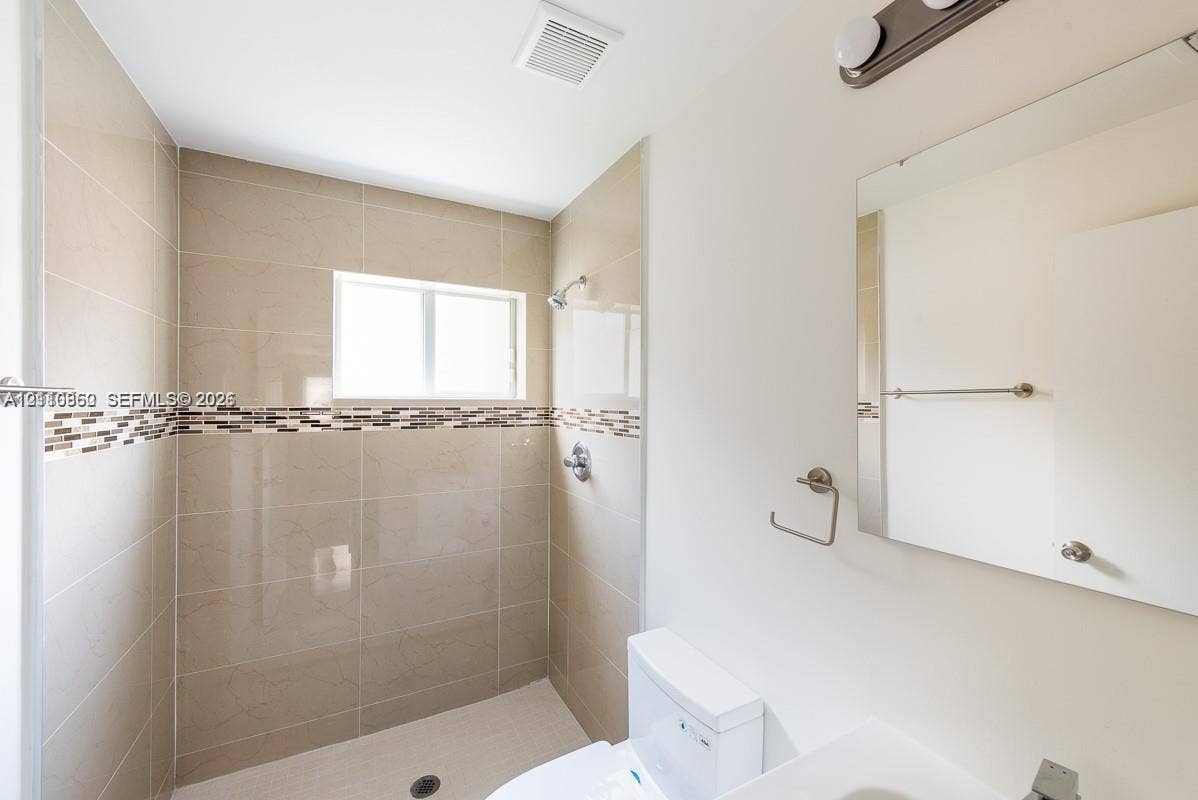 1530 NW 60th Street, Miami, FL 33142 Bathroom featuring a walk-in shower with large-format neutral wall tile, decorative mosaic tile inlay, wall-mounted mirror, and vanity lighting