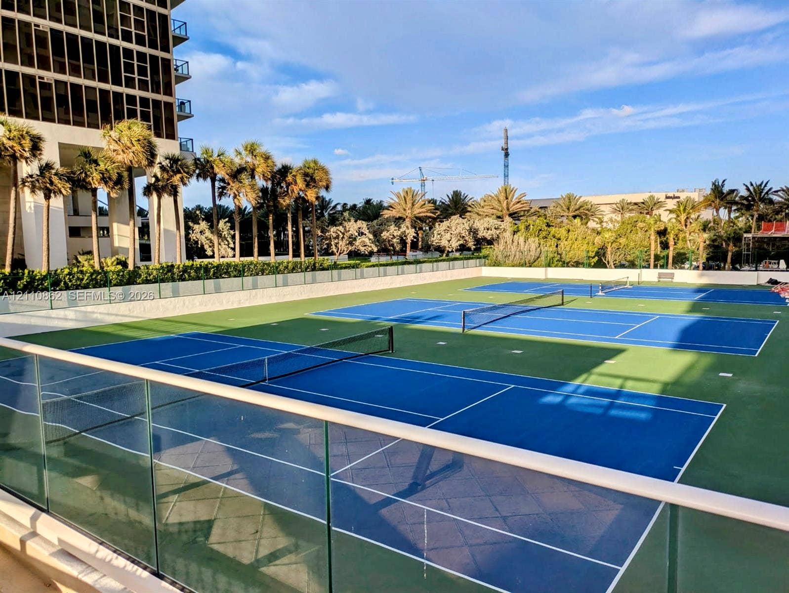 9801 Collins Avenue, #8T, Bal Harbour, Bal Harbour, FL 33154 Multiple professional-grade tennis courts featuring blue and green surfaces, perimeter glass panel fencing, and mature palm tree landscaping