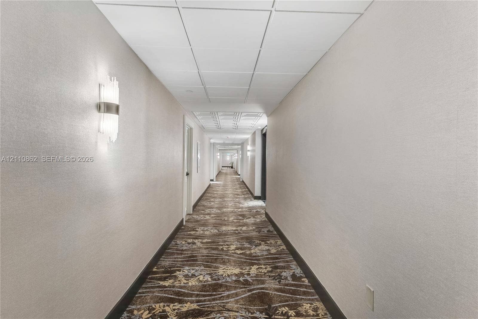 9801 Collins Avenue, #8T, Bal Harbour, Bal Harbour, FL 33154 Hallway featuring neutral wall finishes, textured carpeted flooring, drop ceiling panels, and brushed metal wall-mounted light fixtures