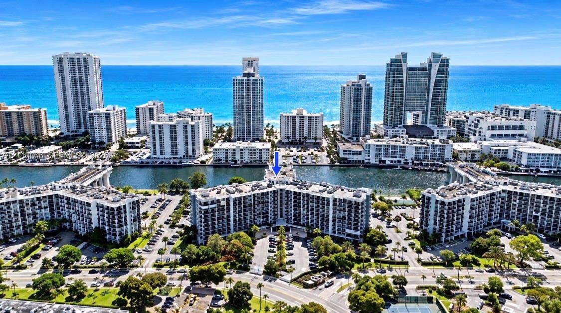 Aerial perspective of a multi-story residential complex situated along a waterway, featuring proximity to coastal waters, landscaped grounds, and paved parking areas