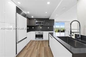 Kitchen featuring high-gloss cabinetry, stainless steel appliances, dark stone-surface countertops, a deep basin sink with a pull-down sprayer faucet, and wood-finish flooring