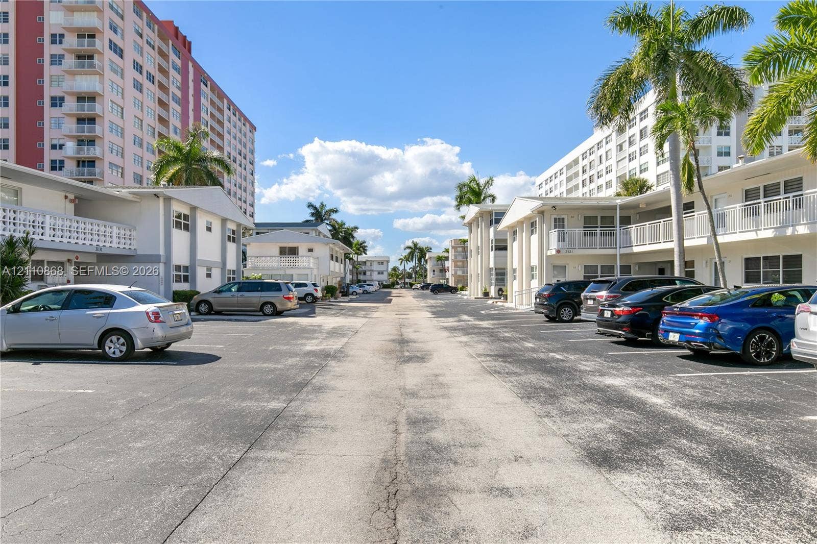 3121 S Ocean Drive, #209, Hallandale Beach, FL 33009 Asphalt parking area featuring multi-story residential buildings, white exterior facades, horizontal balcony railings, and palm trees