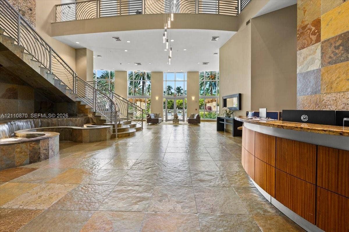 1755 E Hallandale Beach Boulevard, #MEZ5E, Hallandale Beach, FL 33009 Grand lobby featuring dual-height ceilings, stone tile flooring, a curved wood-panel reception desk, a curved stone staircase with metal railing, and floor-to-ceiling windows