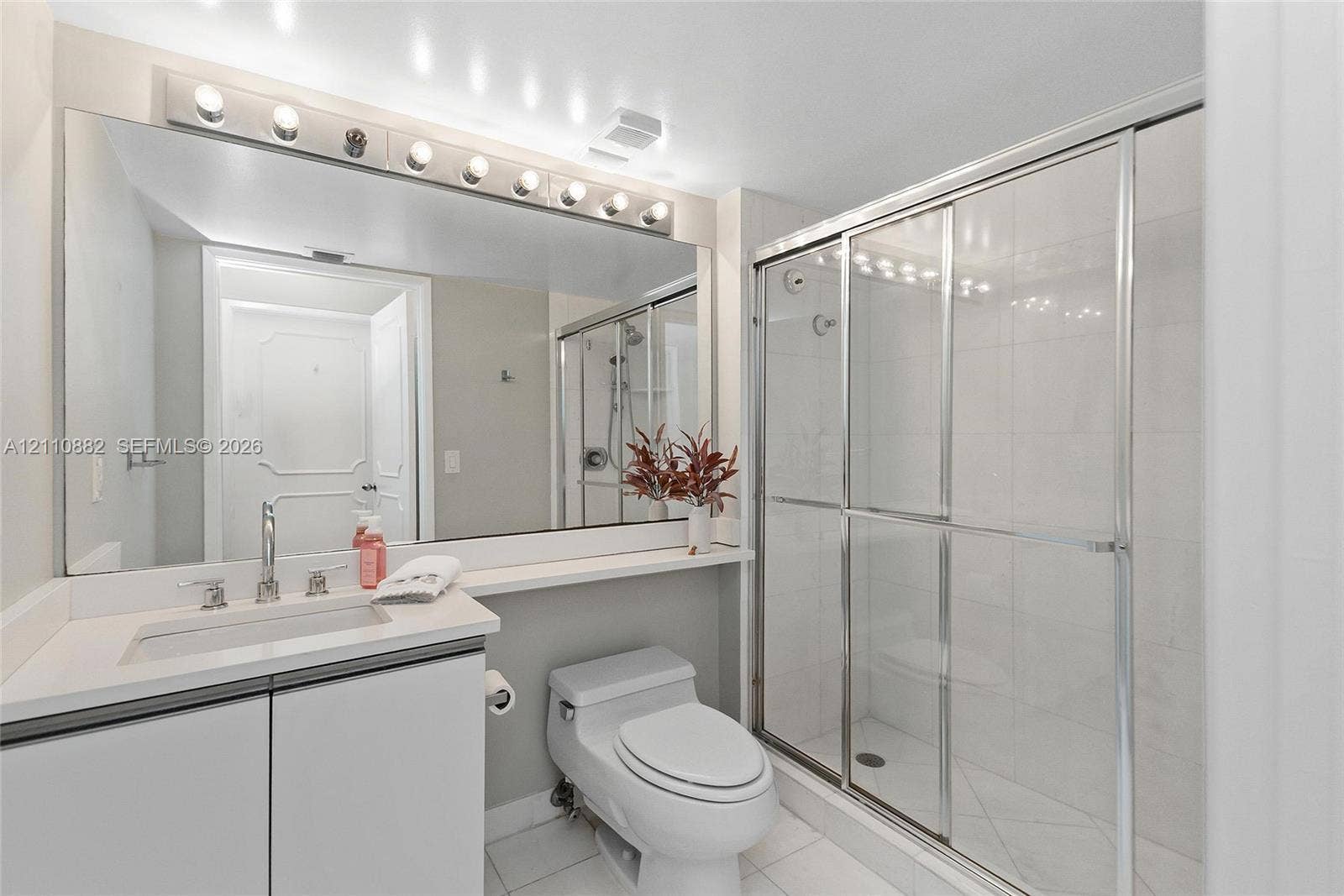 Bathroom featuring a vanity with a white countertop and rectangular undermount sink, expansive wall-to-wall mirror, multi-bulb overhead vanity lighting, and a walk-in shower with a glass sliding door and tiled surround