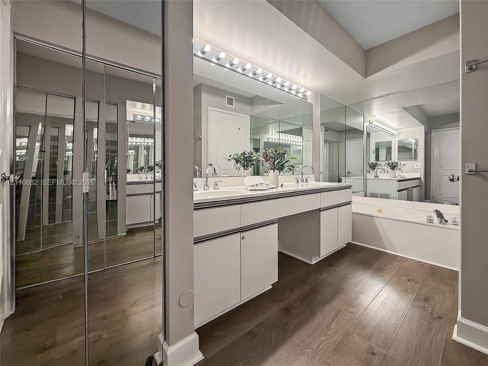 Double vanity featuring white cabinetry, wood-finish flooring, full-wall mirrors, integrated lighting, and a deep soaking tub