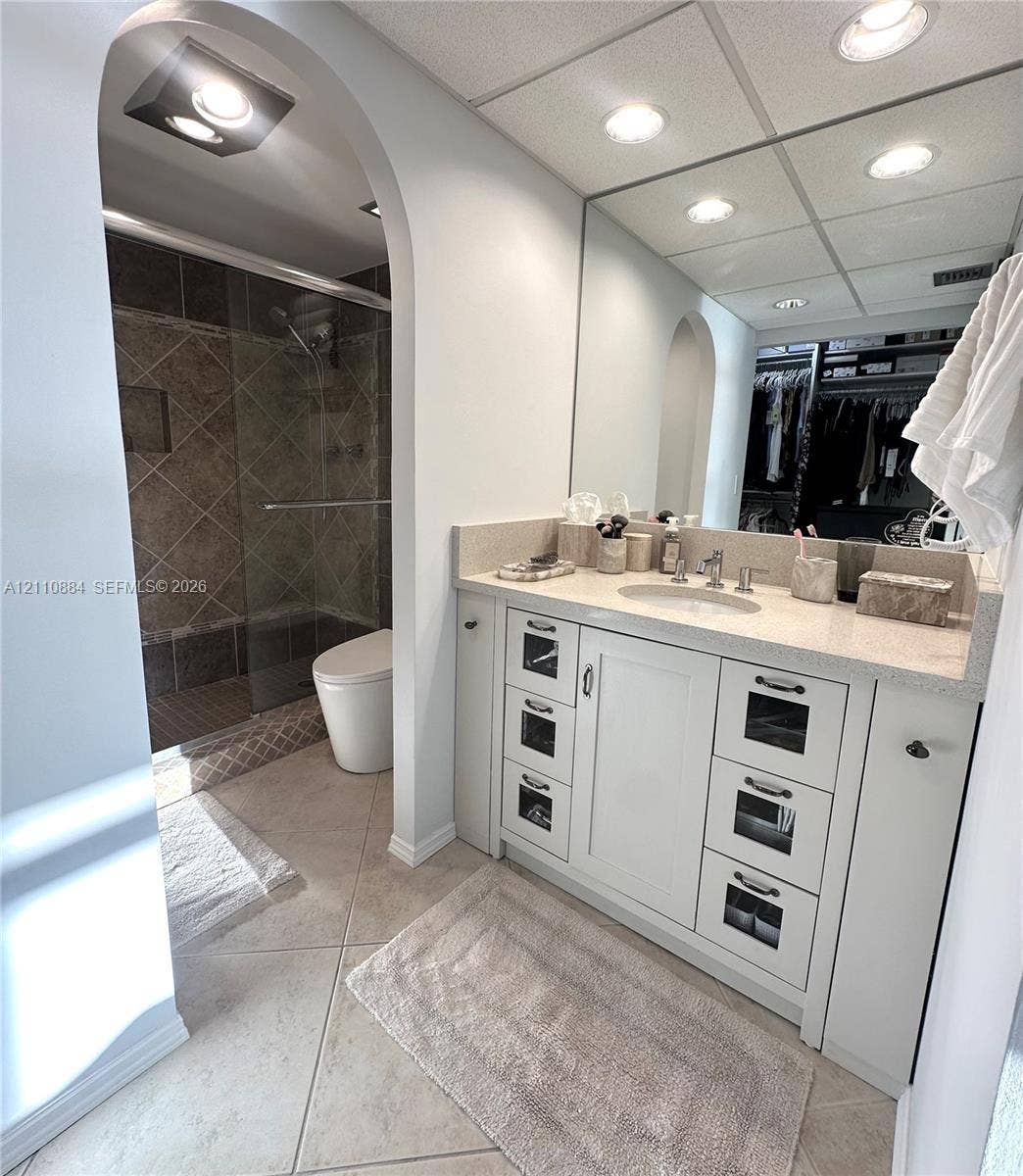 400 Diplomat Parkway, #805, Hallandale Beach, FL 33009 Bathroom featuring a vanity with shaker-style cabinetry, glass-front drawer accents, and a stone-composite countertop with an integrated basin, complemented by an arched doorway, a walk-in shower with tiled surround, and ceramic tile flooring. New ultra-modern style toilet.