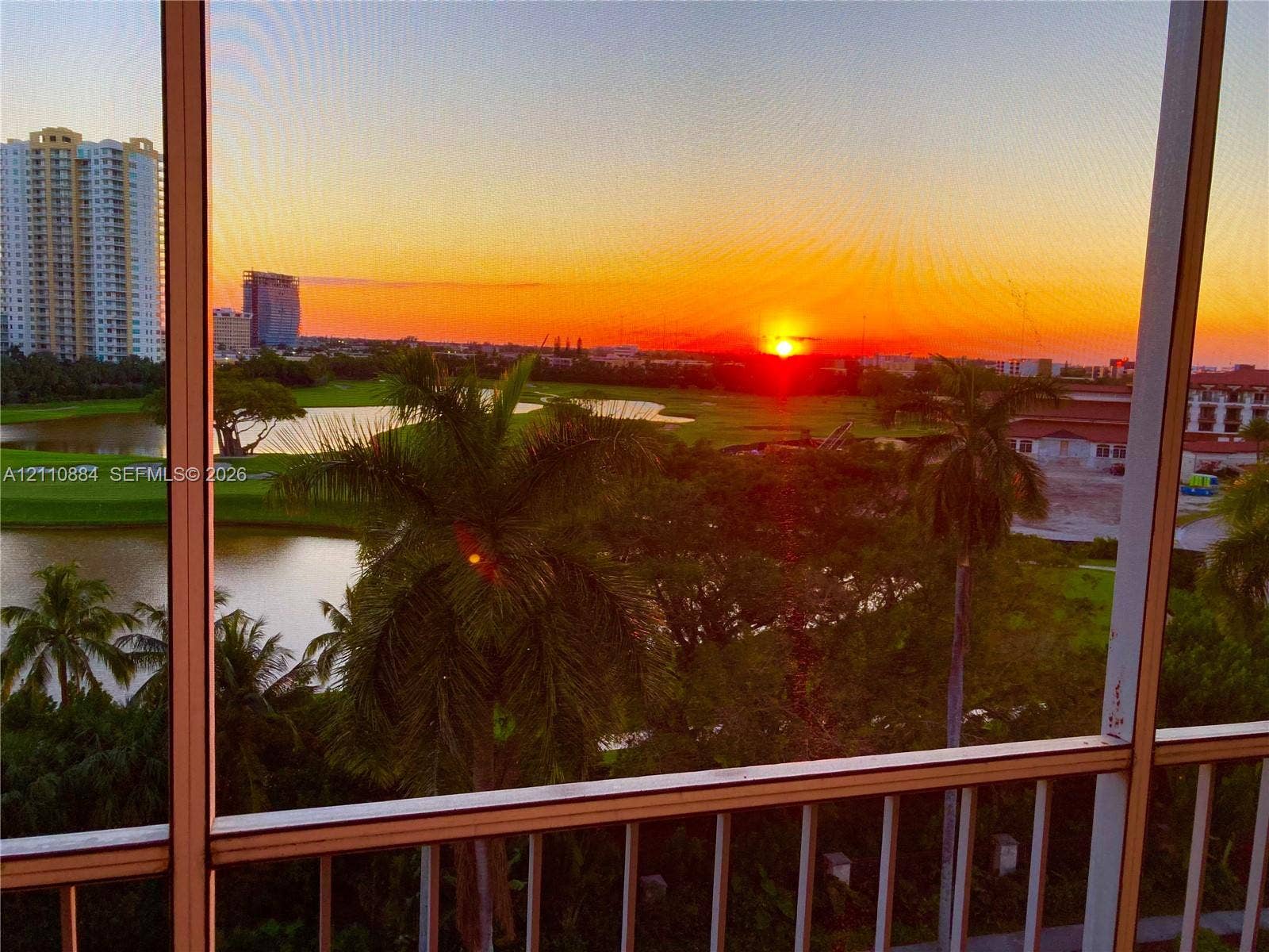 400 Diplomat Parkway, #805, Hallandale Beach, FL 33009 Screened balcony featuring metal railings and a panorama of palm trees, water features, and expansive greenery at sunset