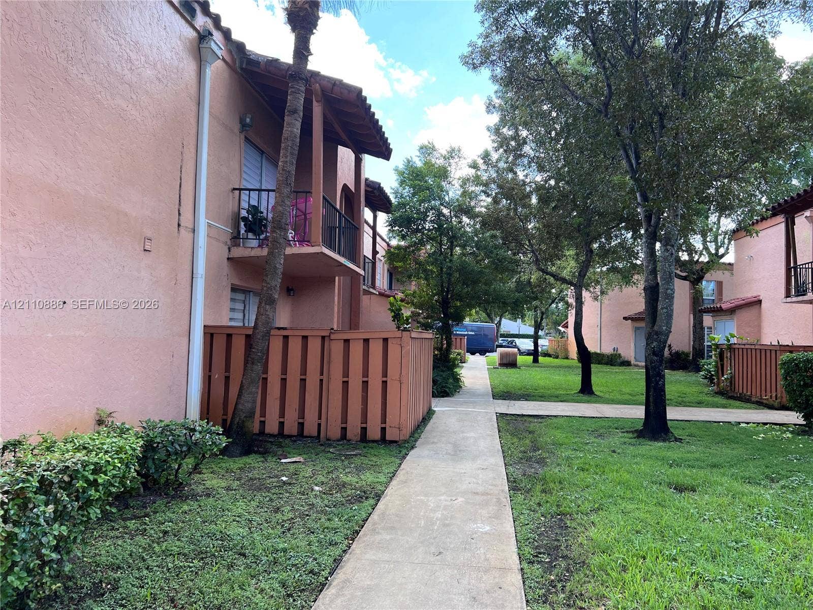 7101 Miami Lakes Drive, #Q12, Miami Lakes, Miami Lakes, FL 33014 Stucco exterior featuring second-story balconies, concrete walkways, and perimeter wood fencing, situated amidst landscaped grounds with mature trees