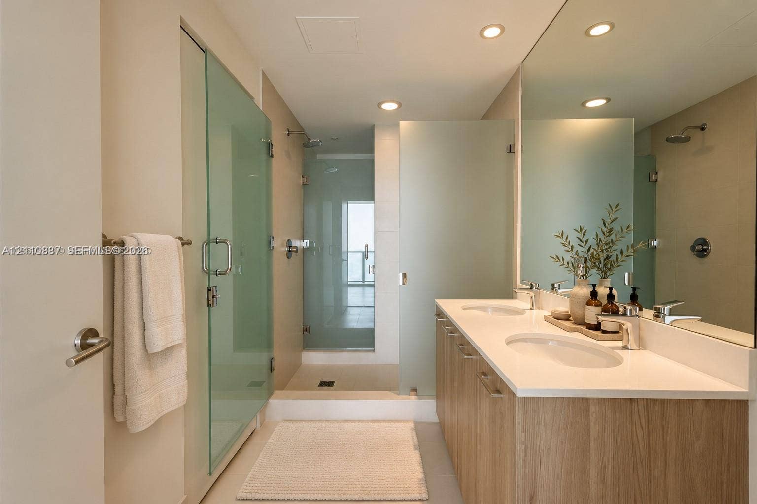 501 NE 31st Street, #4002, Miami, FL 33137 Dual-sink vanity with a solid-surface countertop, wood-finish cabinetry, full-width wall mirror, and dual glass-enclosed shower stalls with rainfall fixtures