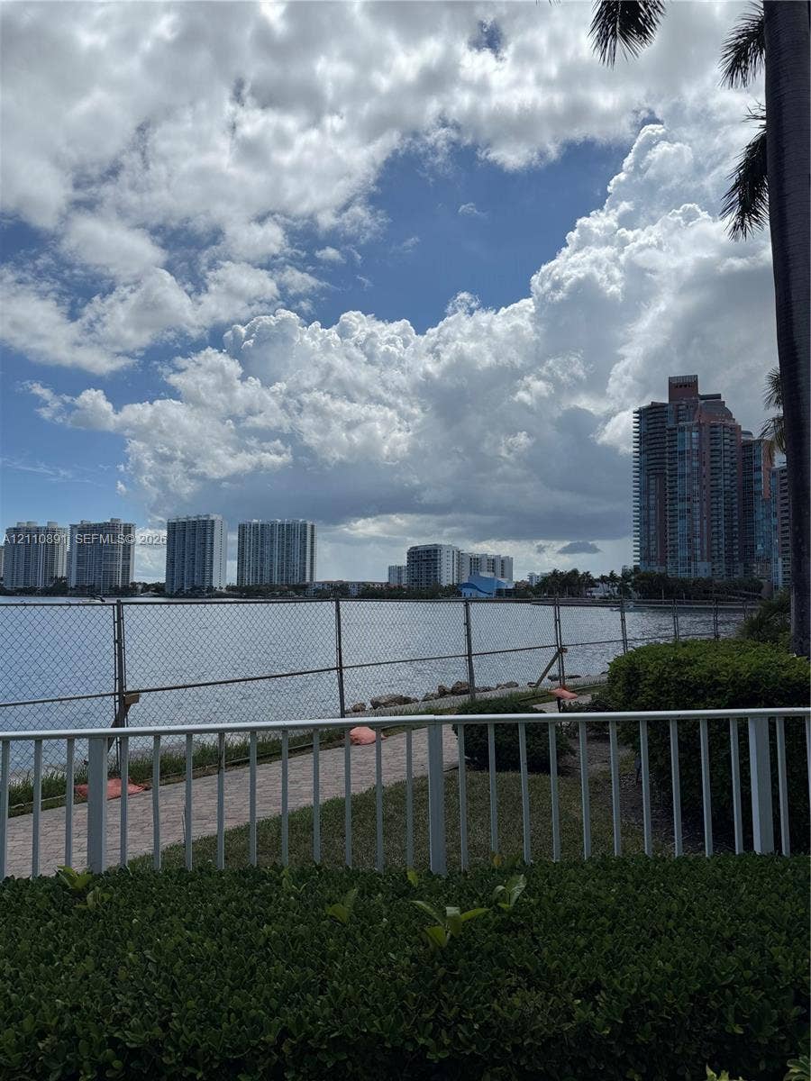 3600 Mystic Pointe Drive, #1611, Aventura, FL 33180 Waterfront grounds featuring a paved walkway, manicured hedging, metal fencing, and perimeter palms