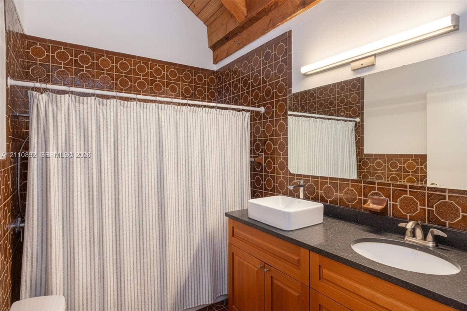 497 N University Drive, #2-7, Plantation, FL 33324 Bathroom featuring geometric-patterned wall tiling, a wood-finish vanity with a dark countertop, a rectangular vessel sink, and an exposed wood beam ceiling