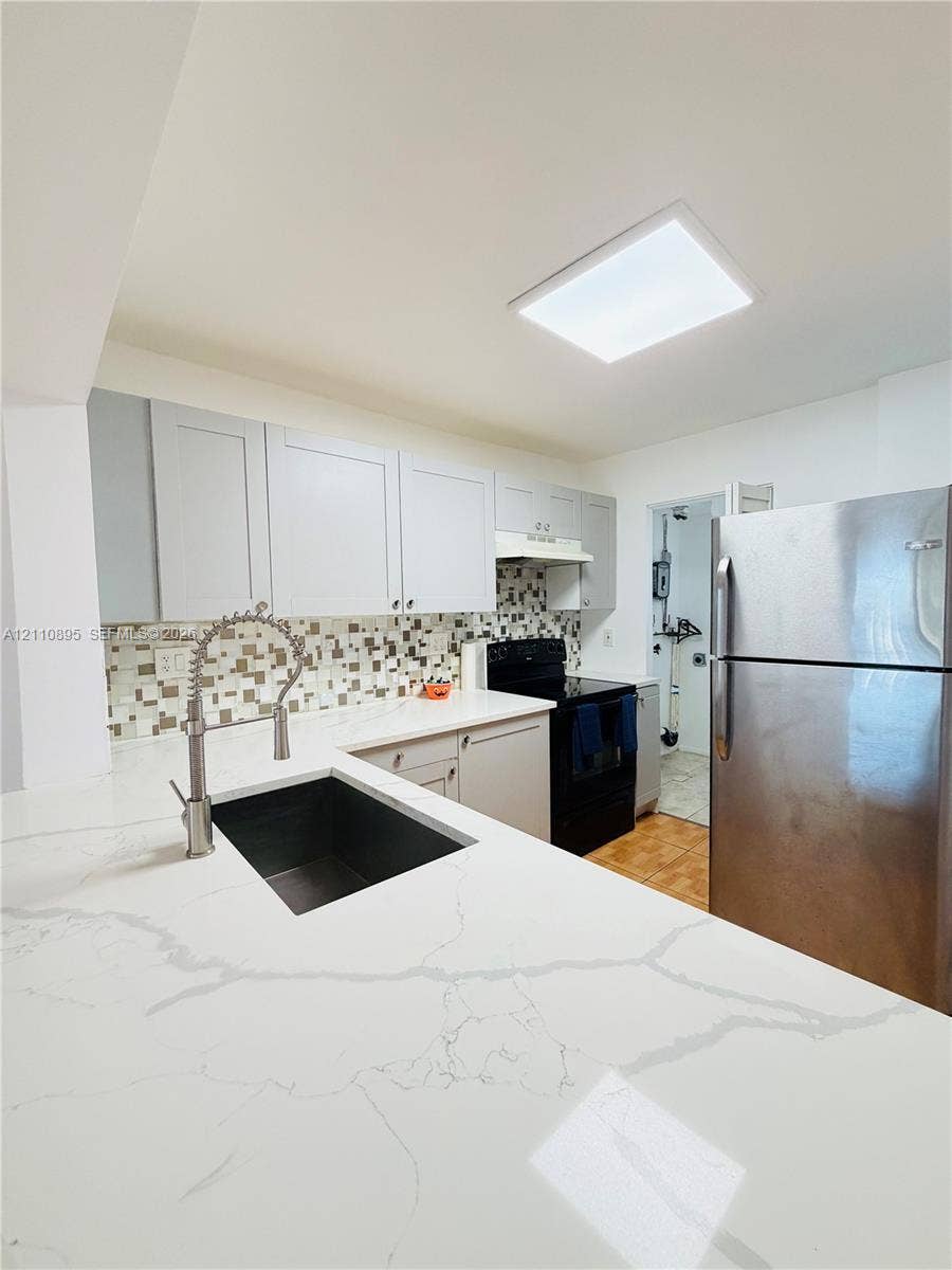 Kitchen featuring stone-patterned countertops, an undermount sink with a gooseneck pull-down faucet, mosaic tile backsplash, shaker-style cabinetry, and stainless steel refrigerator