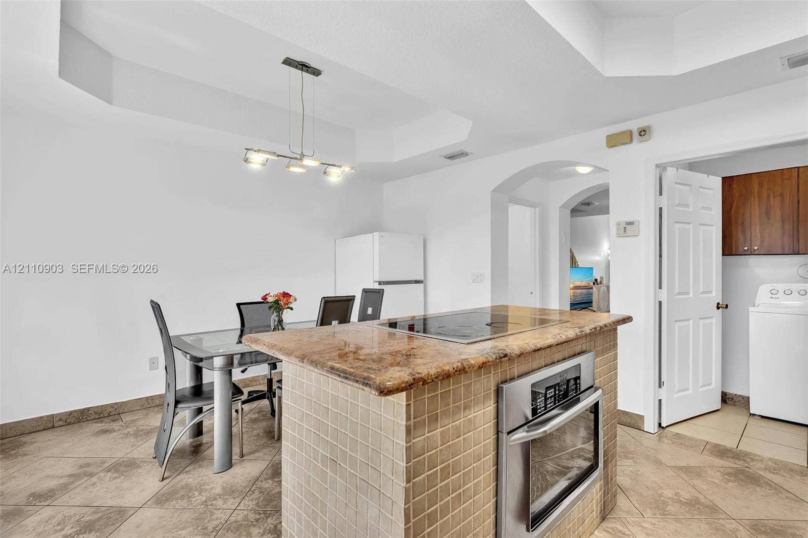 Kitchen island featuring a tiled base, integrated cooktop, and built-in wall oven, complemented by tile flooring, recessed tray ceiling, and a modern multi-light pendant fixture