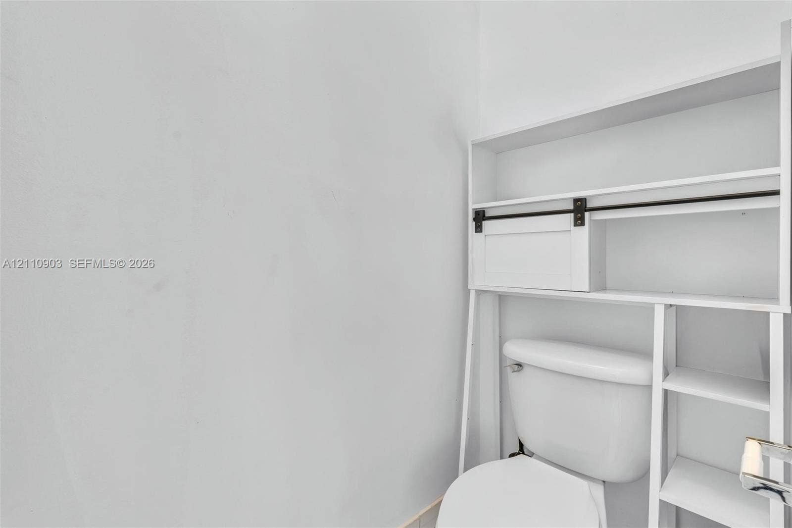 White toilet fixture featuring an over-the-toilet storage unit with white shelving, a sliding cabinet door, and black hardware