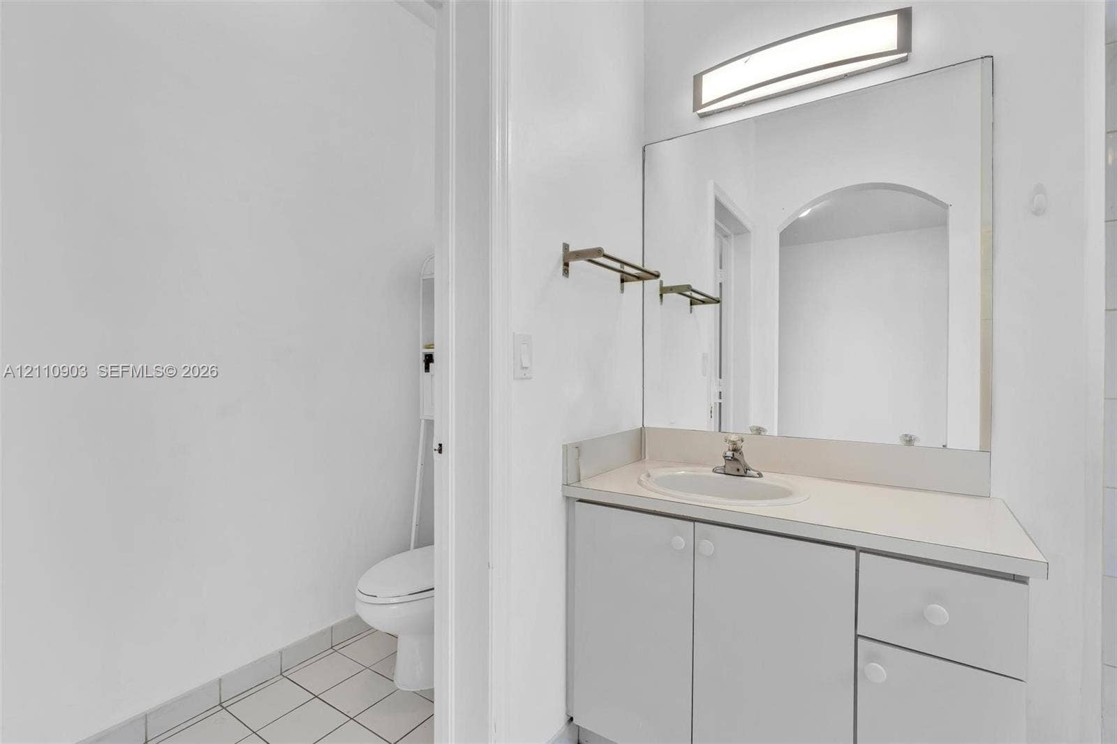 Bathroom vanity featuring light-toned cabinetry, an integrated sink, a full-width mirror, and a wall-mounted light fixture, paired with tile flooring and a standard-height toilet