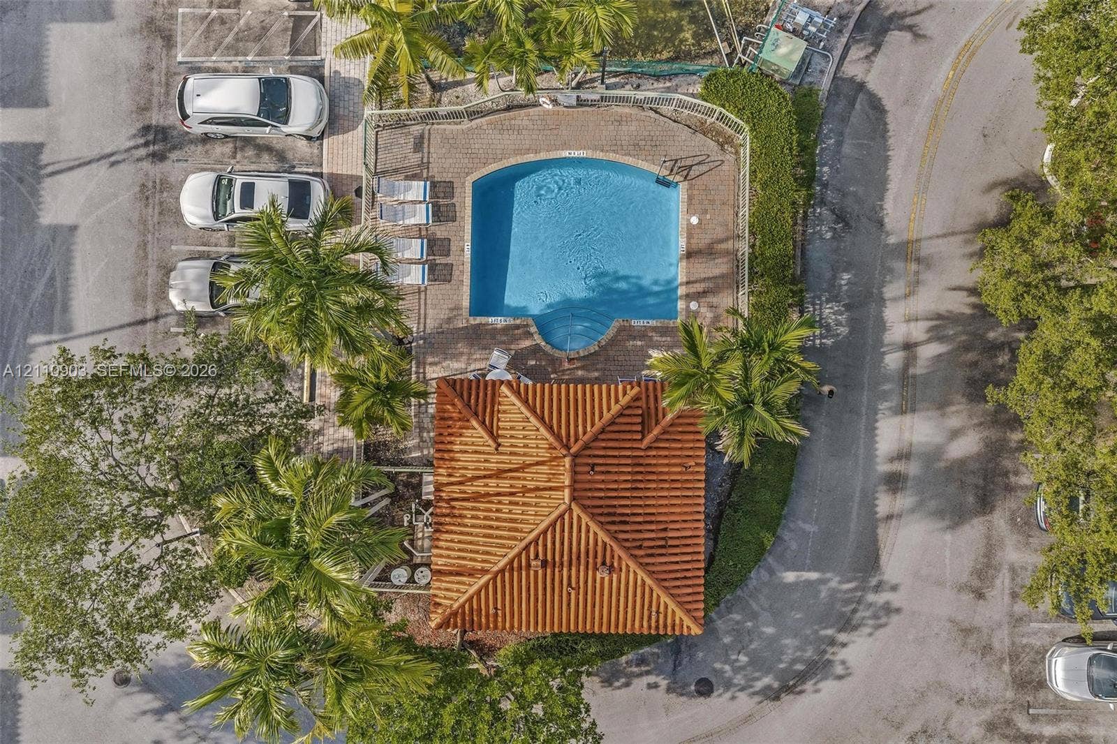 Aerial perspective of a community pool area featuring a curved basin, paver patio, terracotta-style roof structure, and mature palm trees