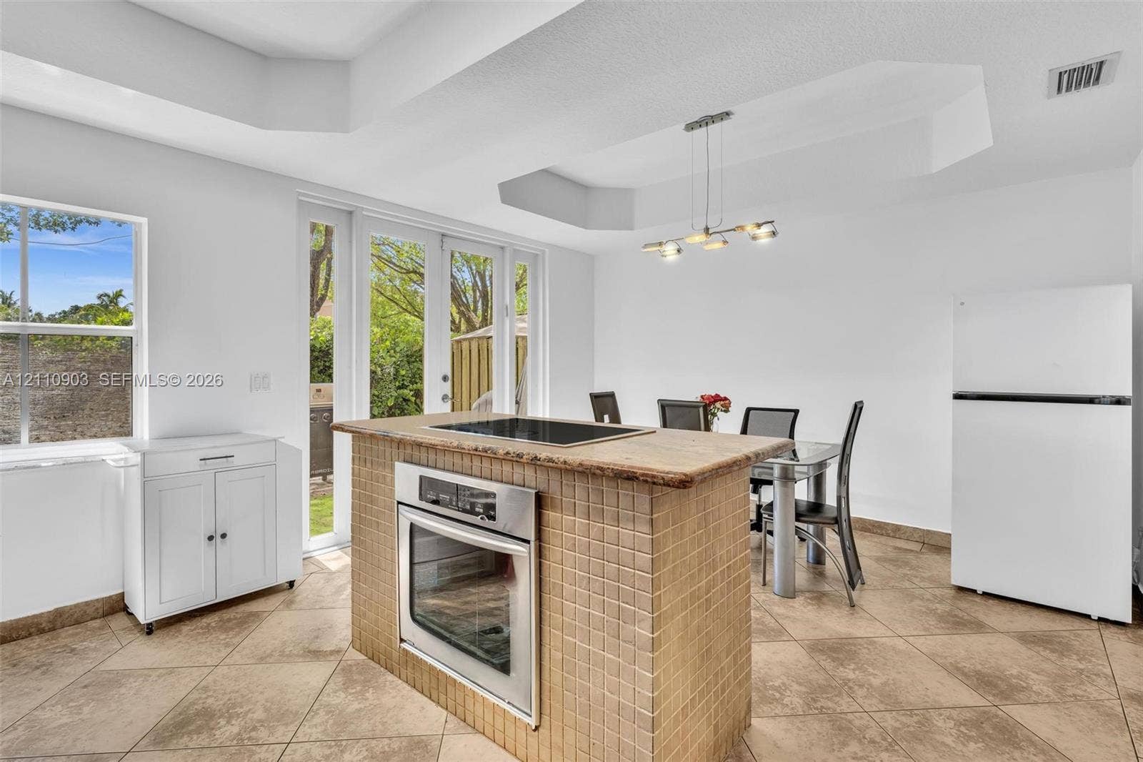 Kitchen island featuring a tiled exterior, integrated cooktop, and built-in stainless steel oven, paired with neutral floor tiling, a tray ceiling, and glass-paneled exterior doors