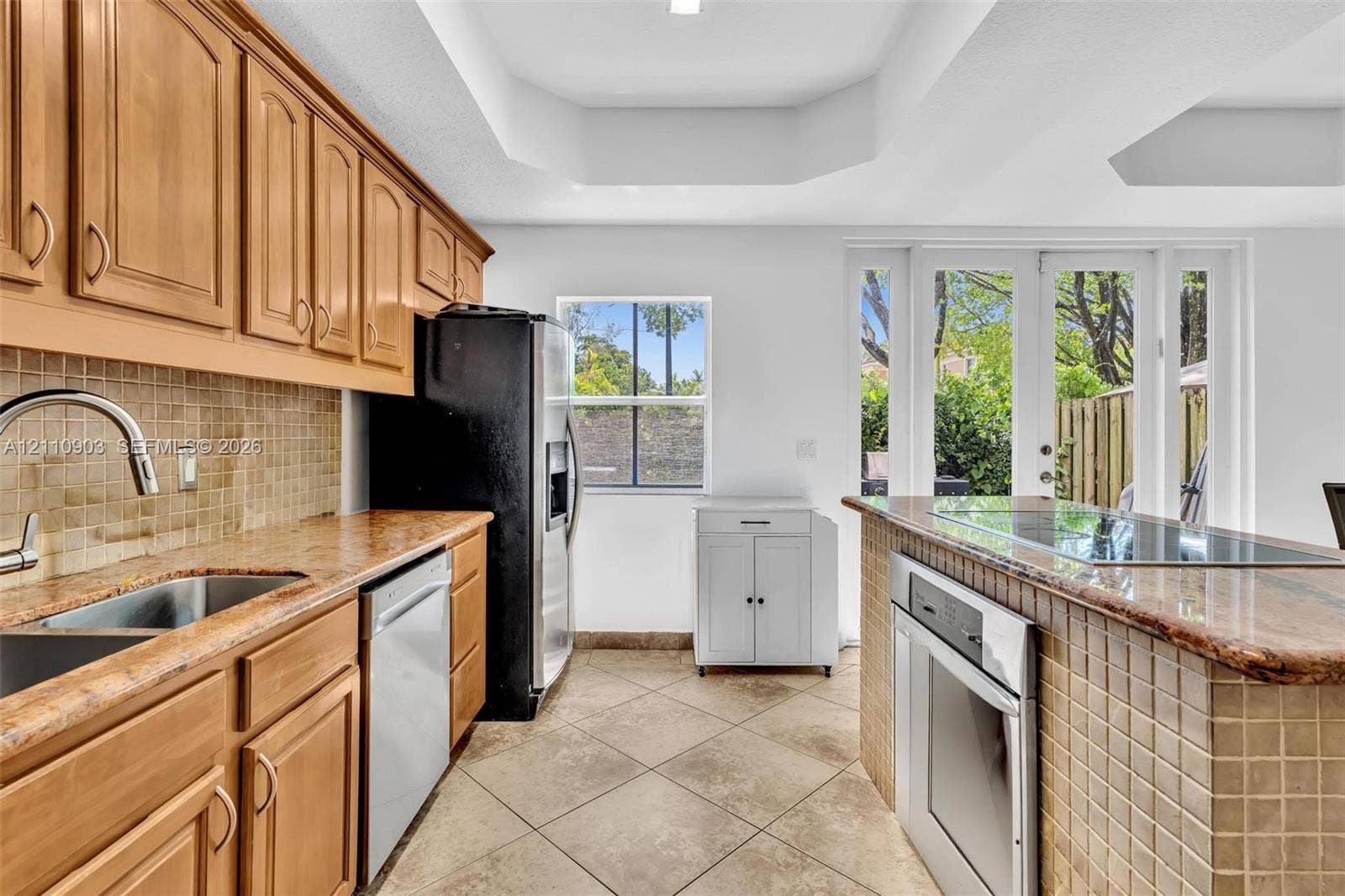 Kitchen featuring honey-toned wood cabinetry, stone countertops, square-tile backsplash, tray ceiling, and French doors